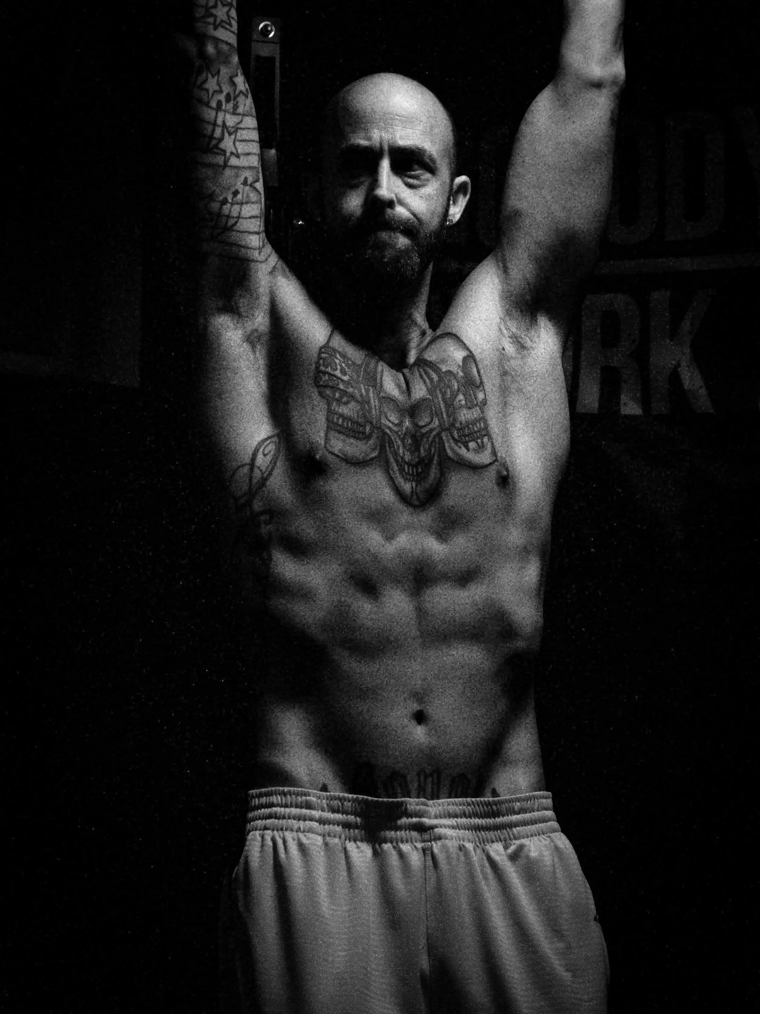 Black and white photo of a muscular, tattooed man with a beard, performing pull-ups. The man is shirtless, showing tattoos on his chest, arms, and abdomen, and is wearing light-colored shorts.