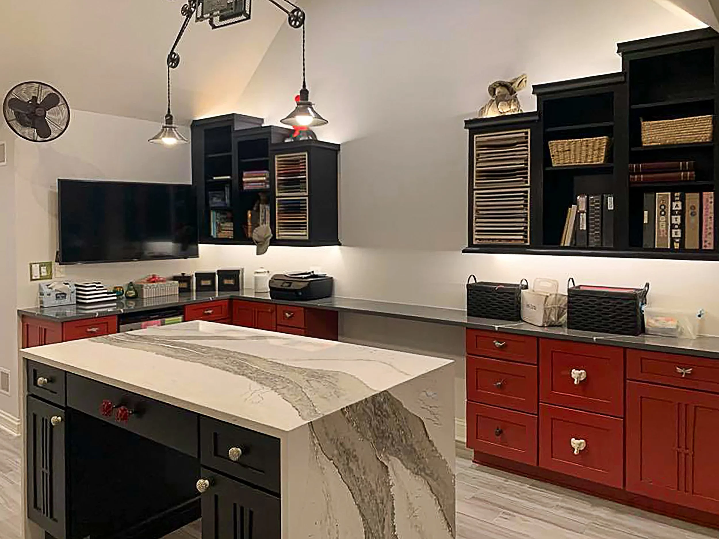 Kitchen with a large, white marble island, black and red cabinetry, open black shelves, a wall-mounted TV, and a wall-mounted fan.