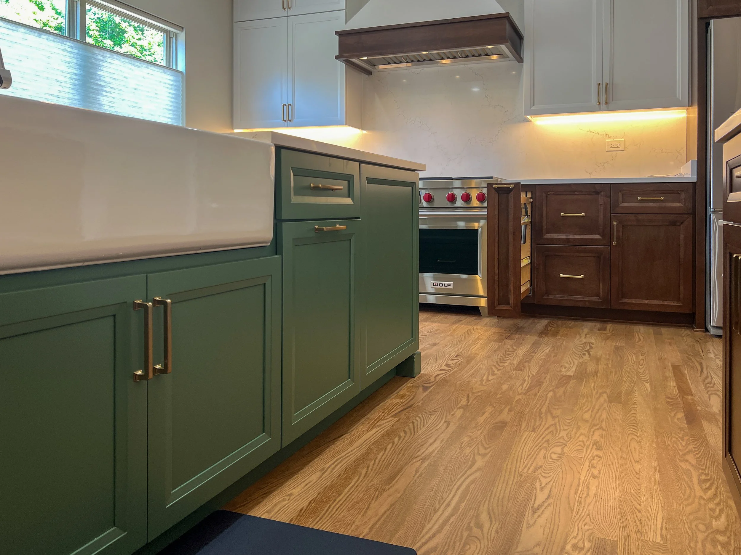 A kitchen with green lower cabinets, brown upper cabinets, white countertops, and a wooden floor. There is a window with sunlight coming through, and a stove with red knobs is visible. The backsplash appears to be a marble or similar material.