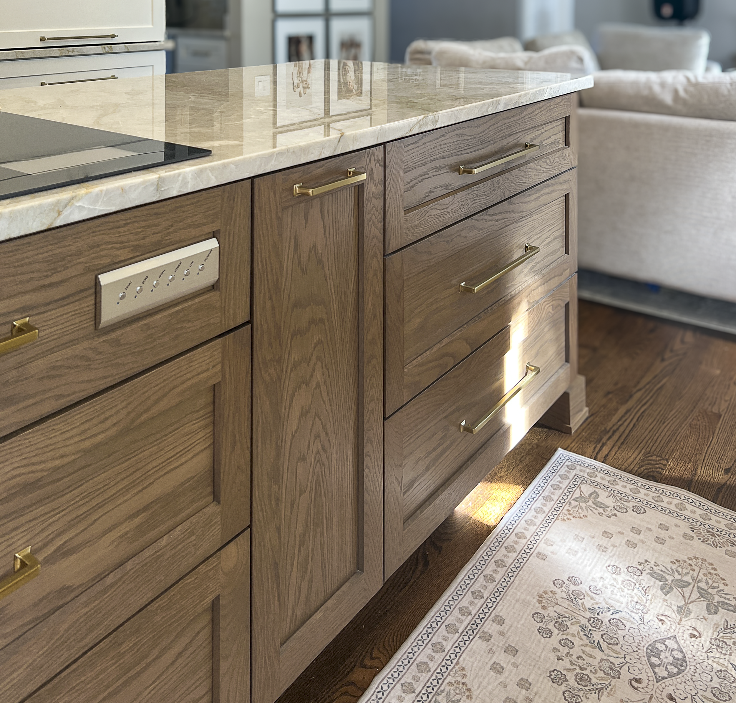 A kitchen island with a marble top, wooden drawers with gold handles, and a built-in oven. In the background, there is a living room with a beige sofa and framed artwork on the wall.