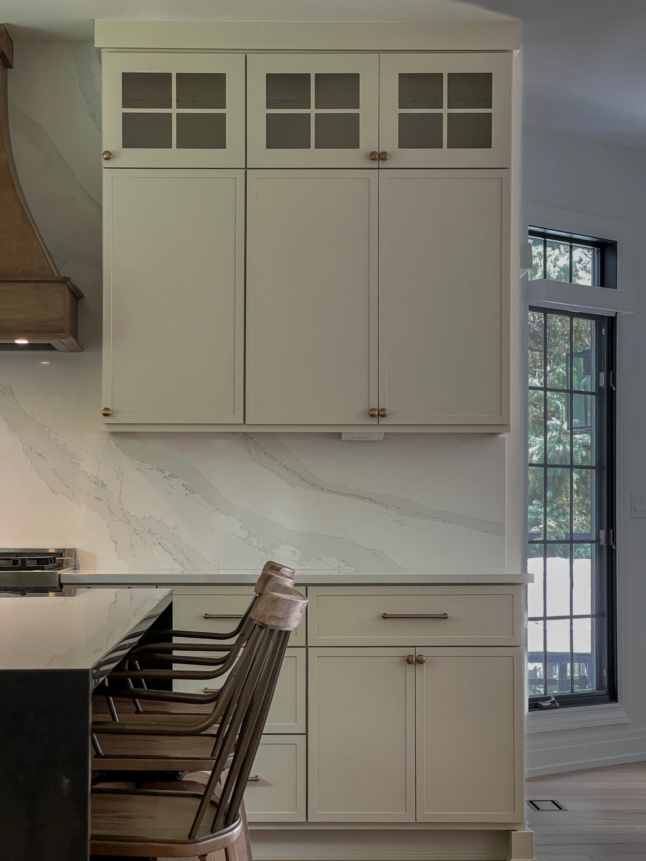 White kitchen cabinet with glass-paneled upper doors and wooden knobs, situated next to a window with black grid framing, part of a modern kitchen with a white marble countertop and chairs in front.