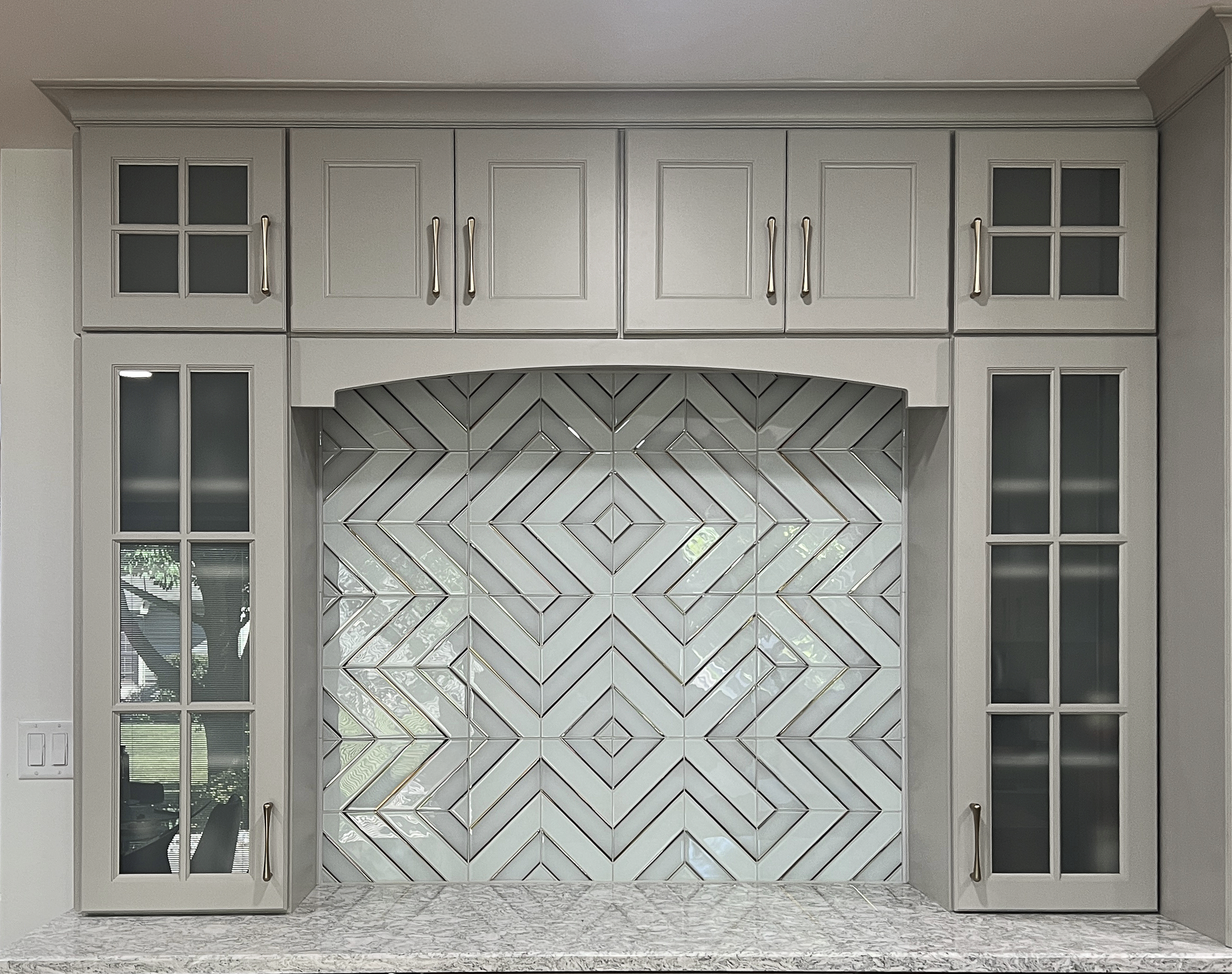 Kitchen cabinet with white doors, gold handles, glass-paneled sections, and a mirrored backsplash with a chevron pattern.