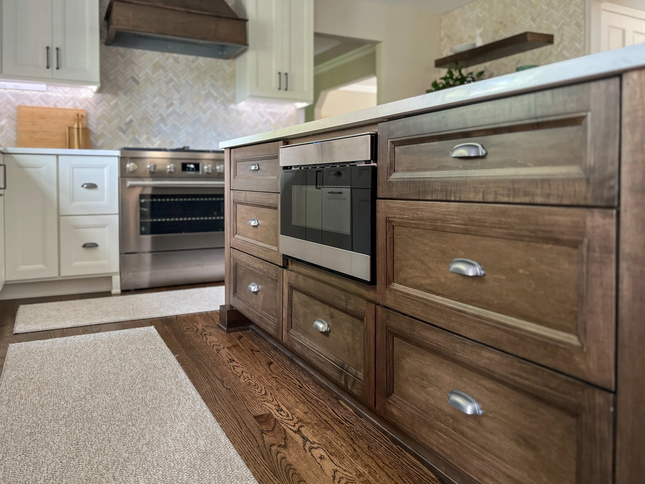 Kitchen with brown wood cabinets and stainless steel appliances, including an oven and a dishwasher, with beige and white backsplash and a beige rug on hardwood floor.