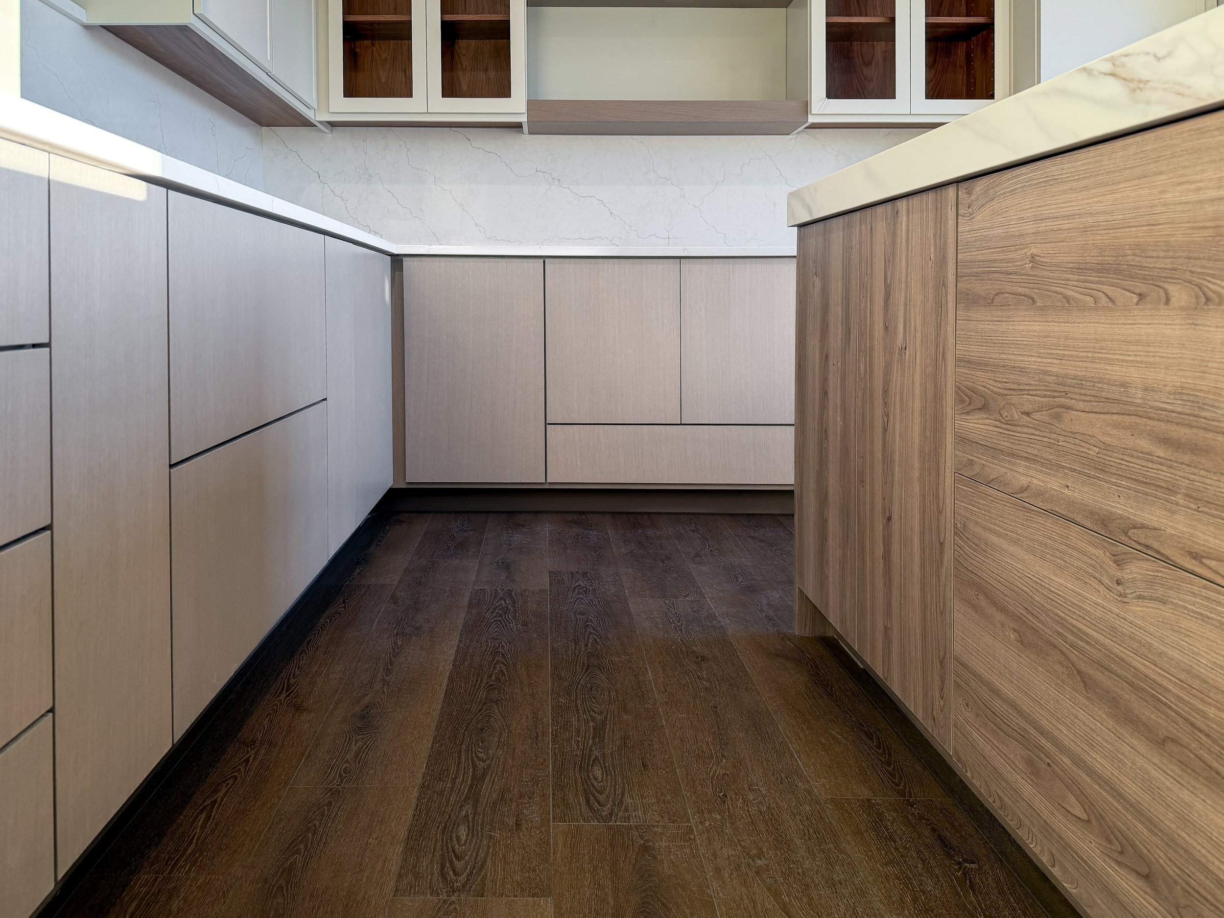 A modern kitchen corner with wooden cabinets, white marble countertops, and a dark wooden floor.