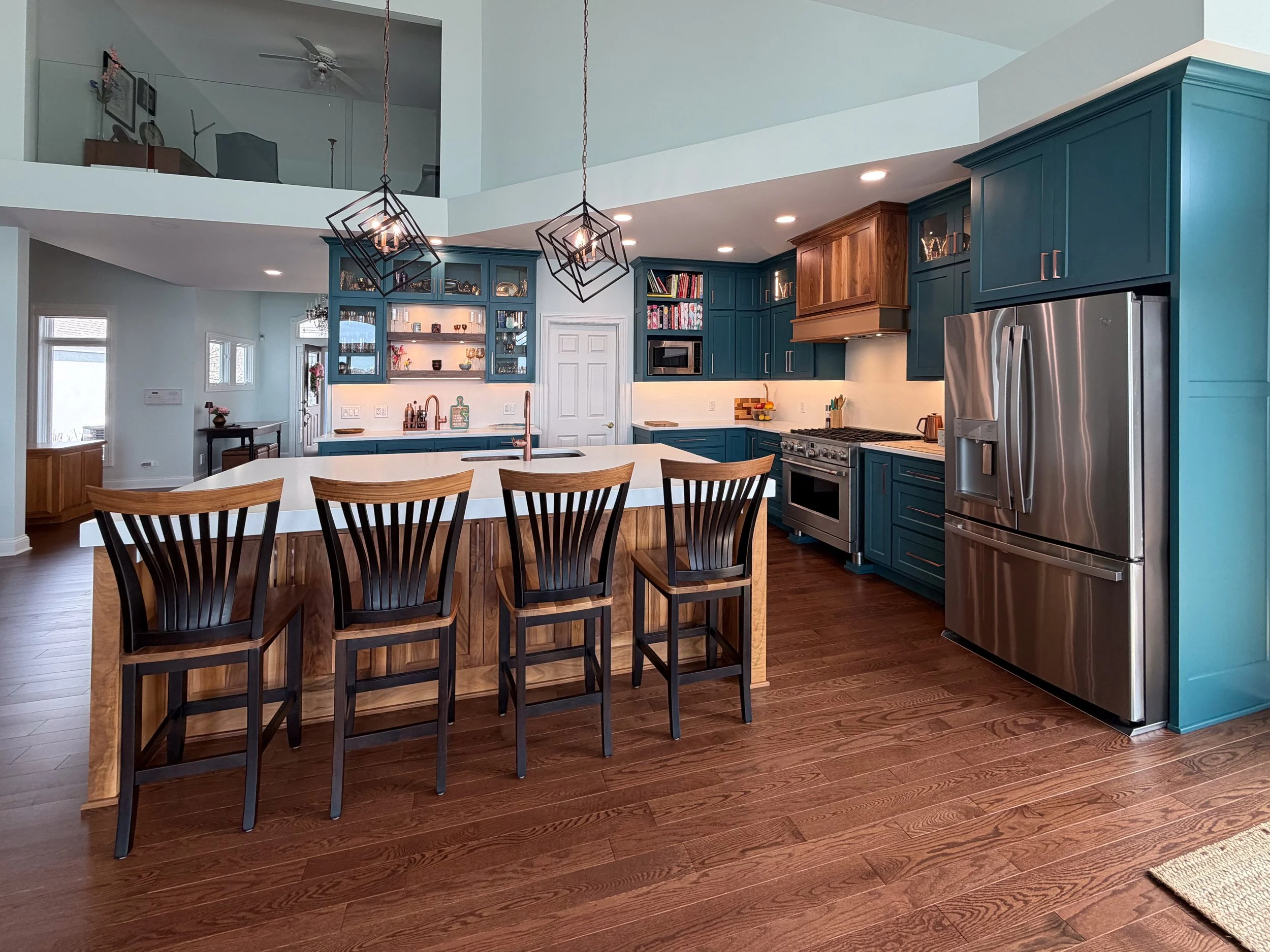 A modern kitchen featuring a large island with wooden base and white countertop, surrounded by black wooden chairs, blue cabinetry, stainless steel appliances, and pendant lights hanging from the ceiling.