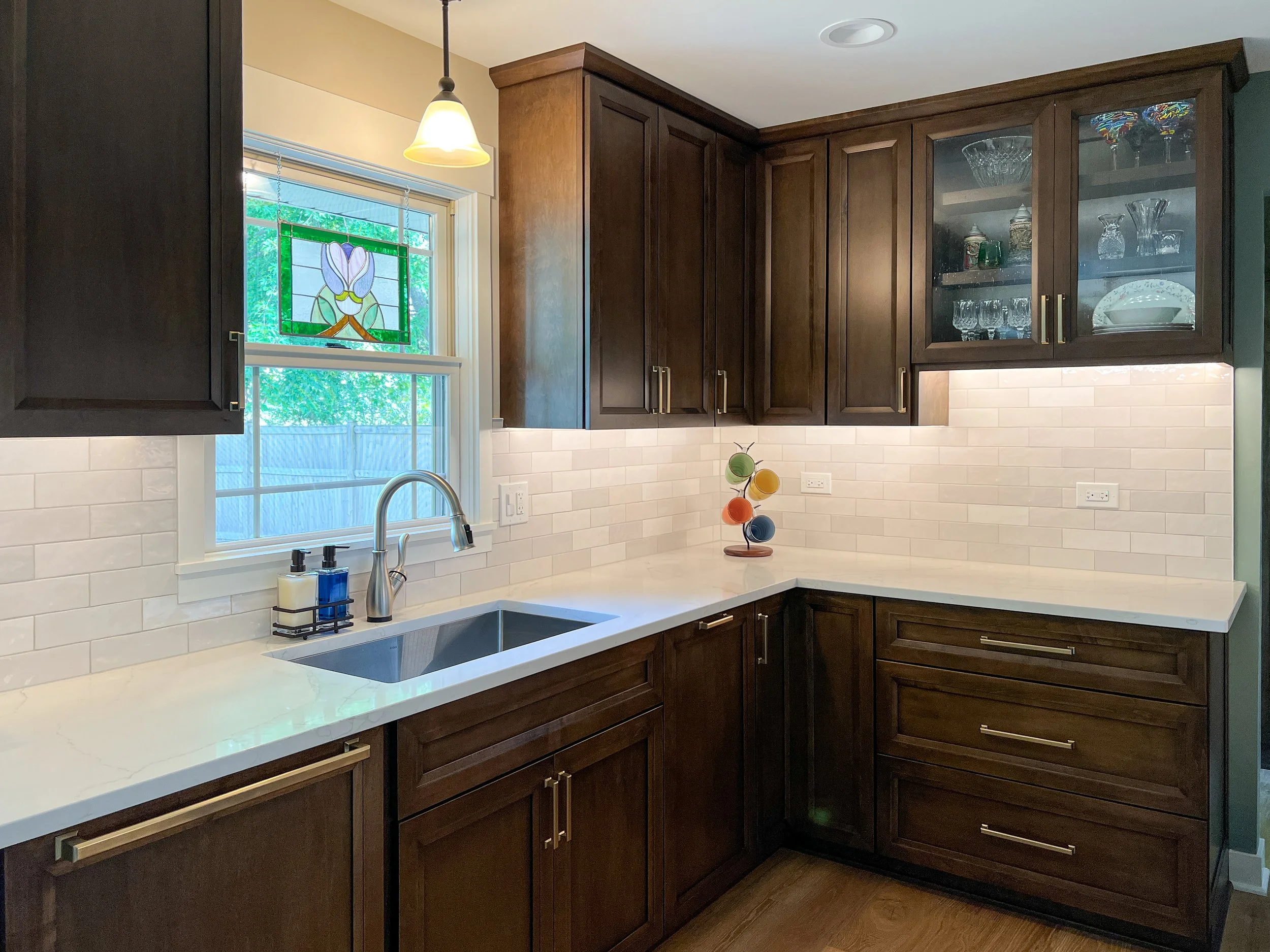 Kitchen with dark wood cabinets, beige countertop, white subway tile backsplash, and a window with a stained glass flower design. There is a small hanging light and a colorful cup tree on the counter.
