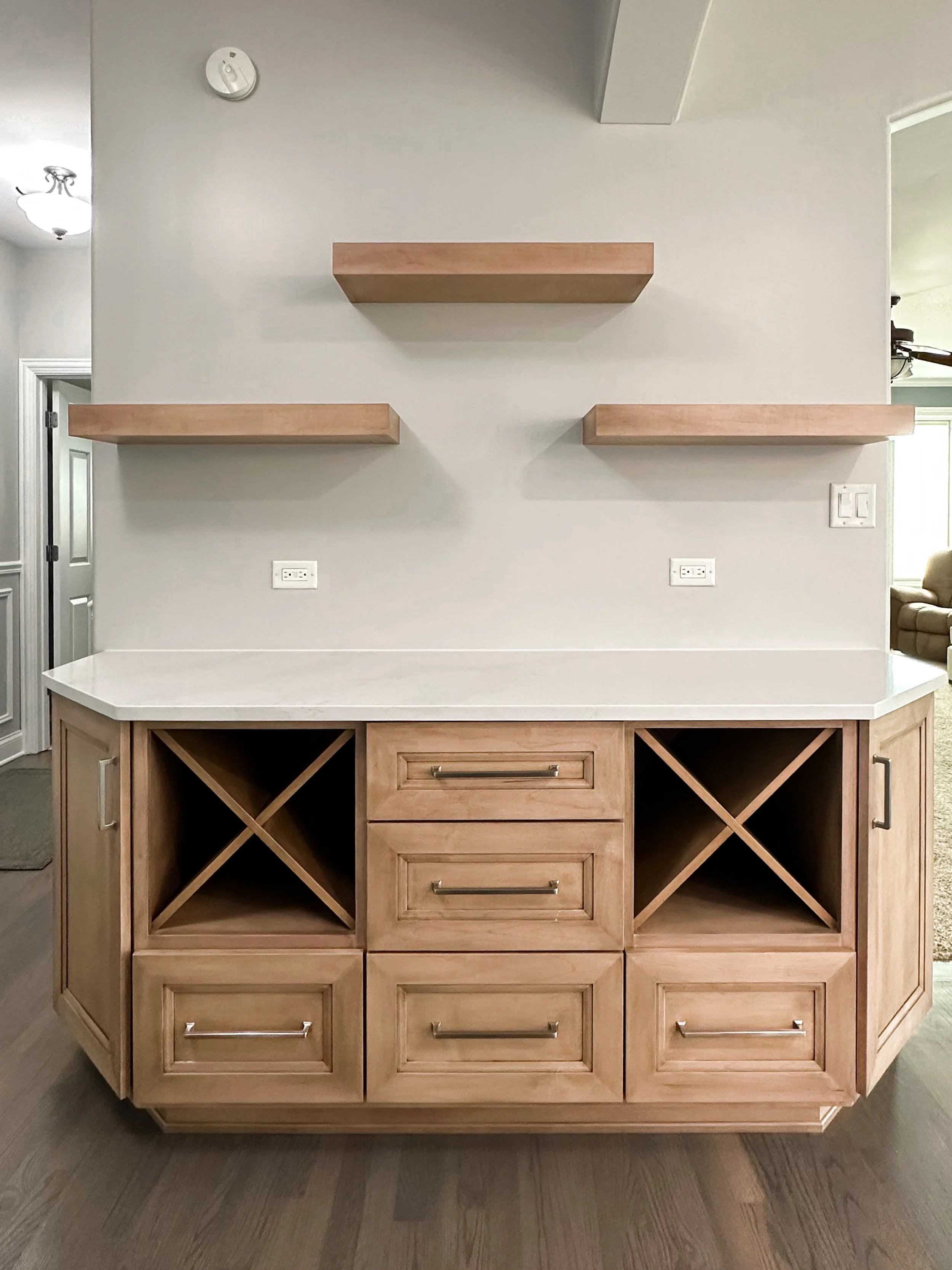 A kitchen island with a cream-colored countertop, wooden drawers and cabinet doors, and lattice wine storage, situated against a light-colored wall with three floating shelves and electrical outlets.