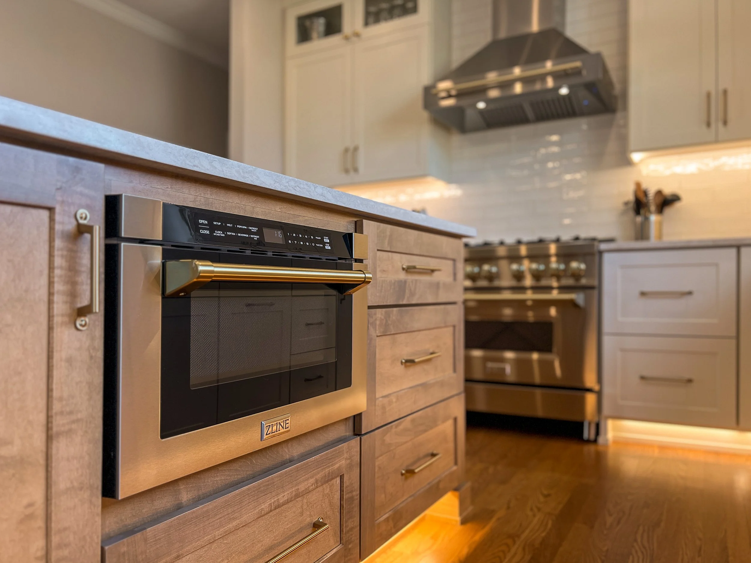Close-up view of a modern stainless steel oven built into a wooden kitchen cabinet, with a gold handle, in a kitchen with beige cabinets, a range hood, and cooking utensils on a countertop in the background.