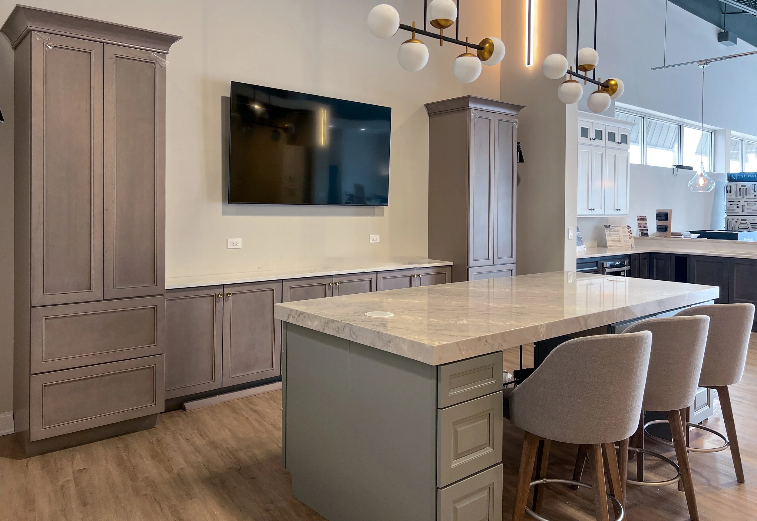 Modern kitchen interior with a large island, surrounded by upholstered chairs, light wood cabinets, a wall-mounted TV, and contemporary lighting fixtures.