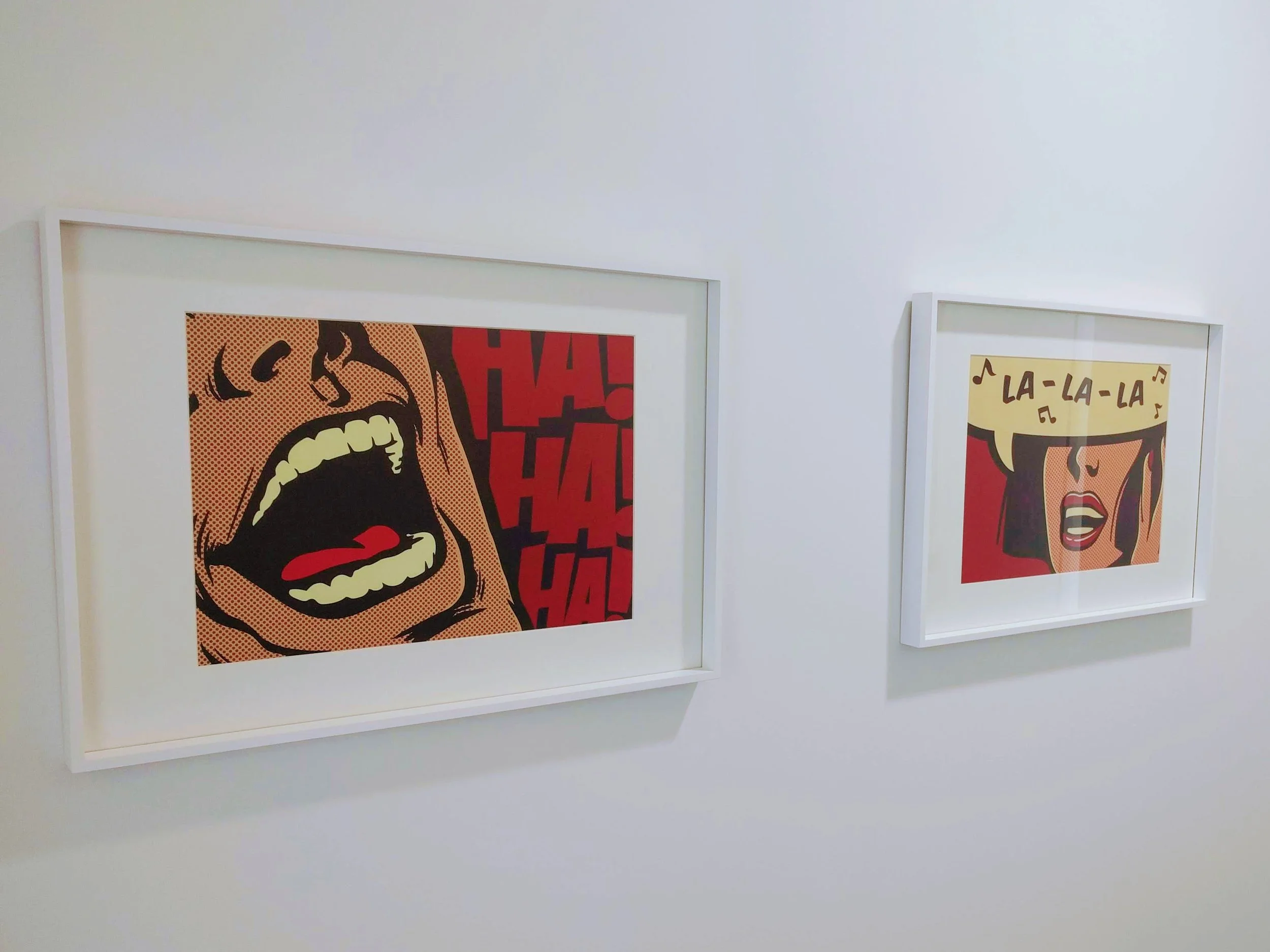 Two framed pop art style prints on a white wall. The left print depicts a person yelling with the word 'HA!' repeatedly in the background. The right print shows a woman singing with a speech bubble saying 'LA-LA-LA' and musical notes.