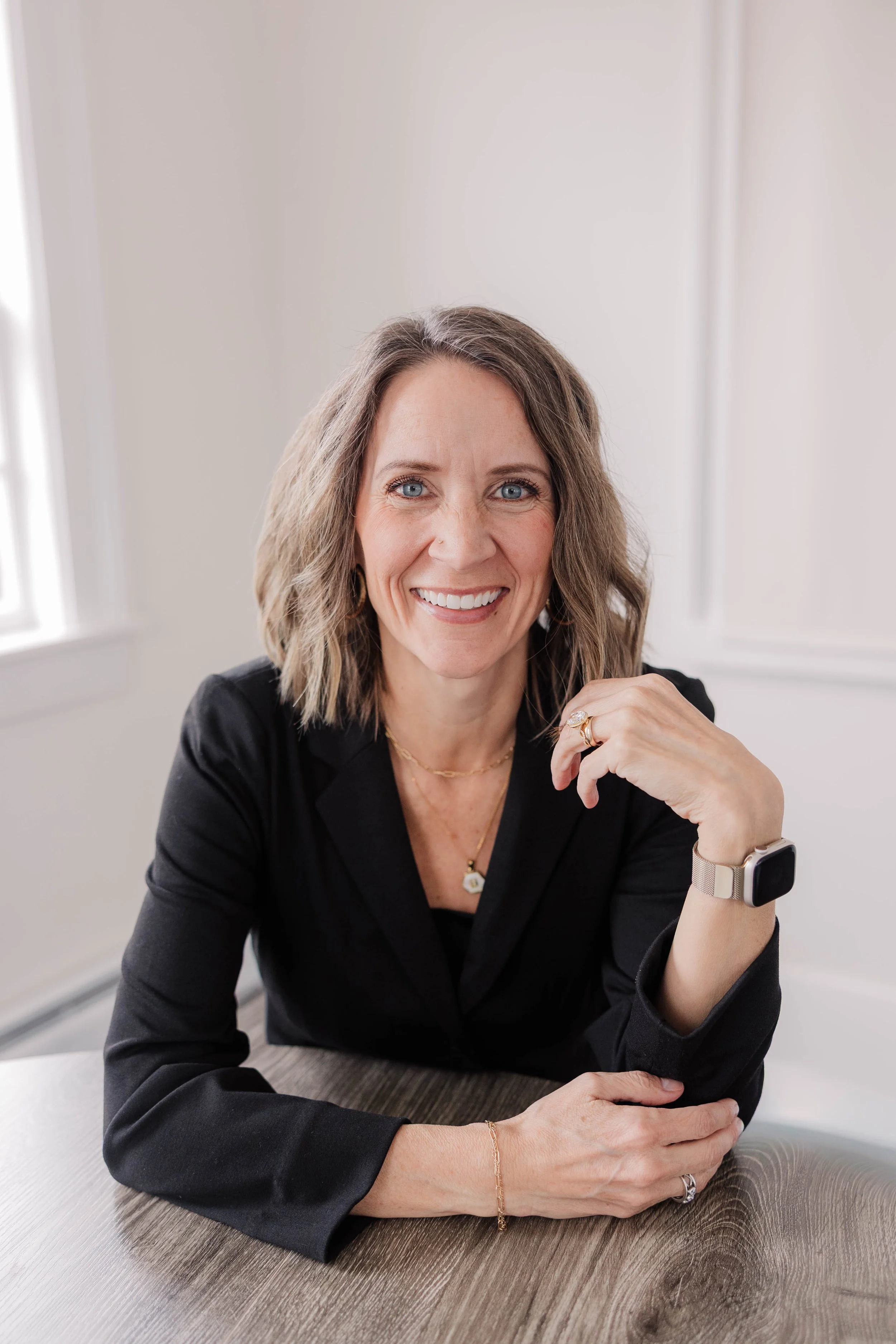 A smiling woman with blue eyes, shoulder-length wavy brown hair, wearing a black blazer, jewelry, and a smartwatch, sitting at a table in a bright room.