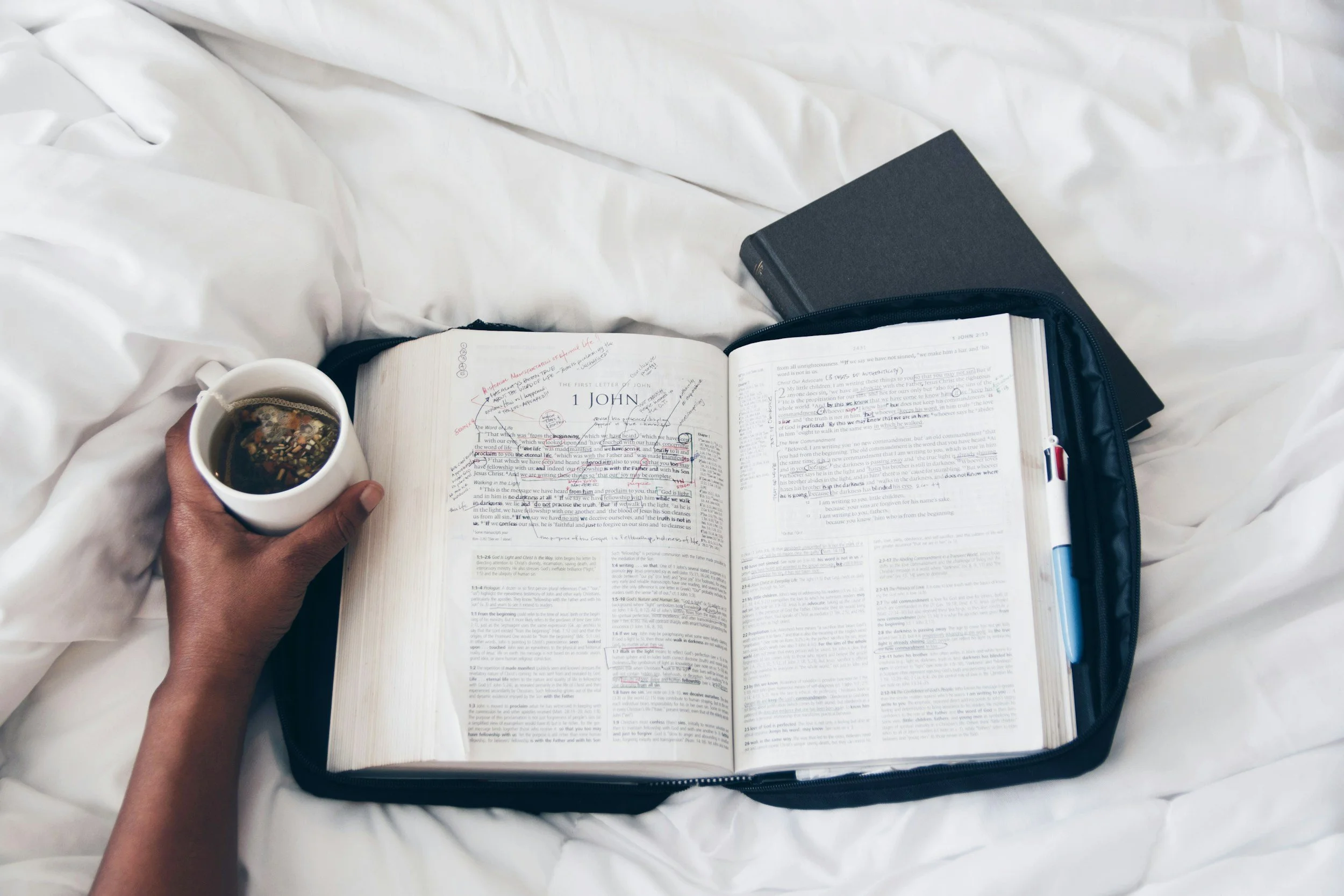 Open Bible with handwritten notes, a hand holding a cup of tea or coffee, and a closed book on a white bed