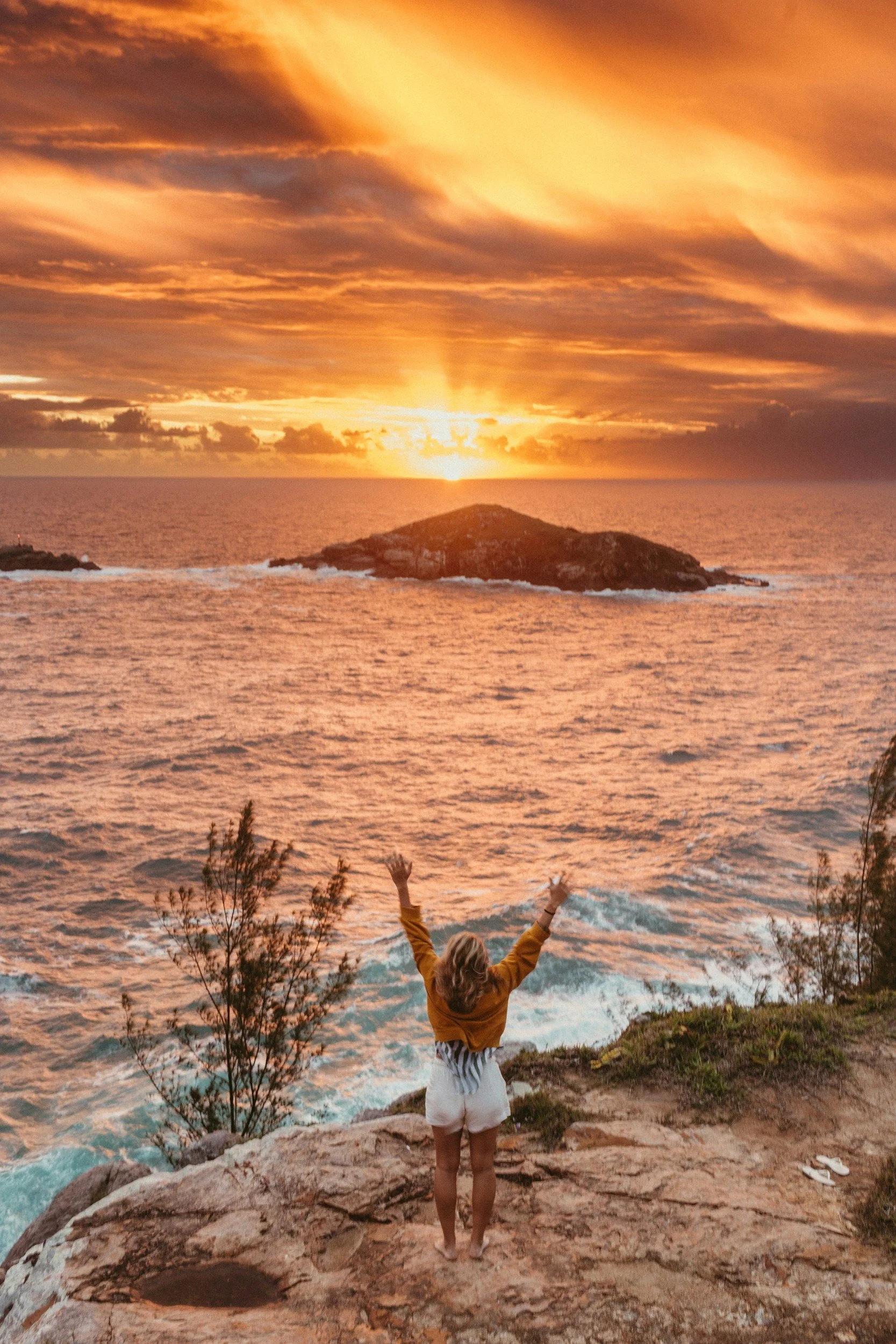 Child standing on rocky ledge with arms raised toward the sunset over ocean, with clouds and small islands in the background.