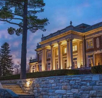 A large, illuminated mansion with classical architecture, columns, and a stone wall in front, surrounded by trees at dusk.