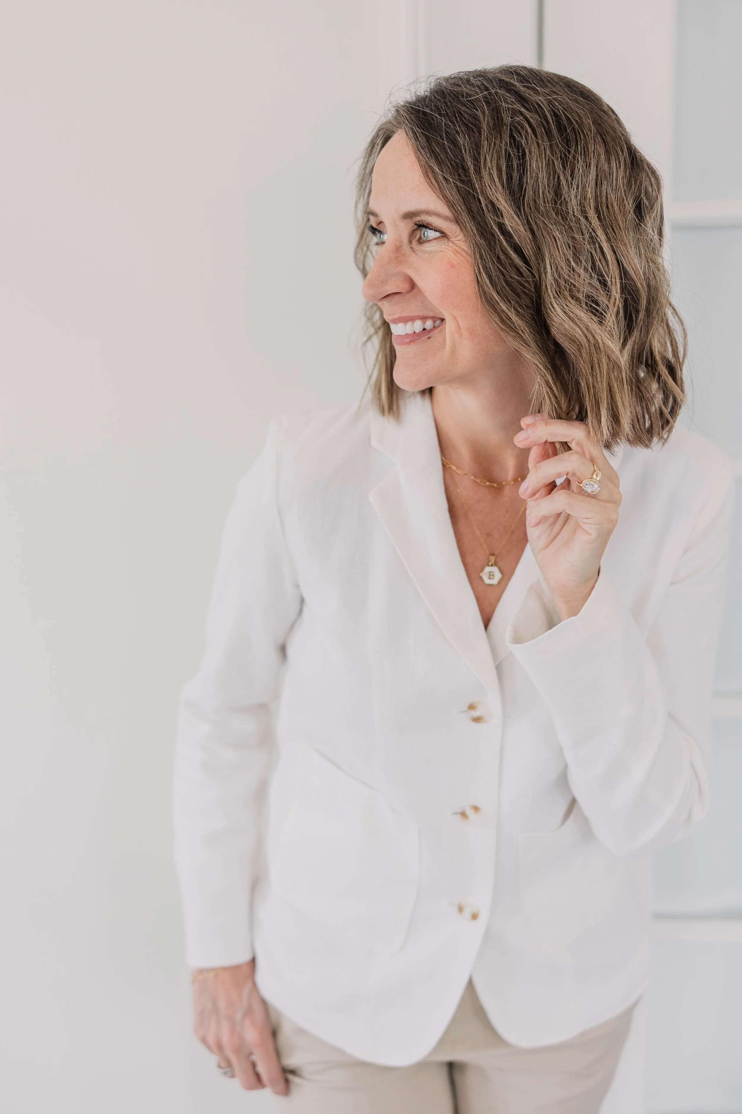 A woman with wavy brown hair and blue eyes smiling while touching her neck, wearing a white button-up shirt and layered gold necklaces, standing against a plain light background.