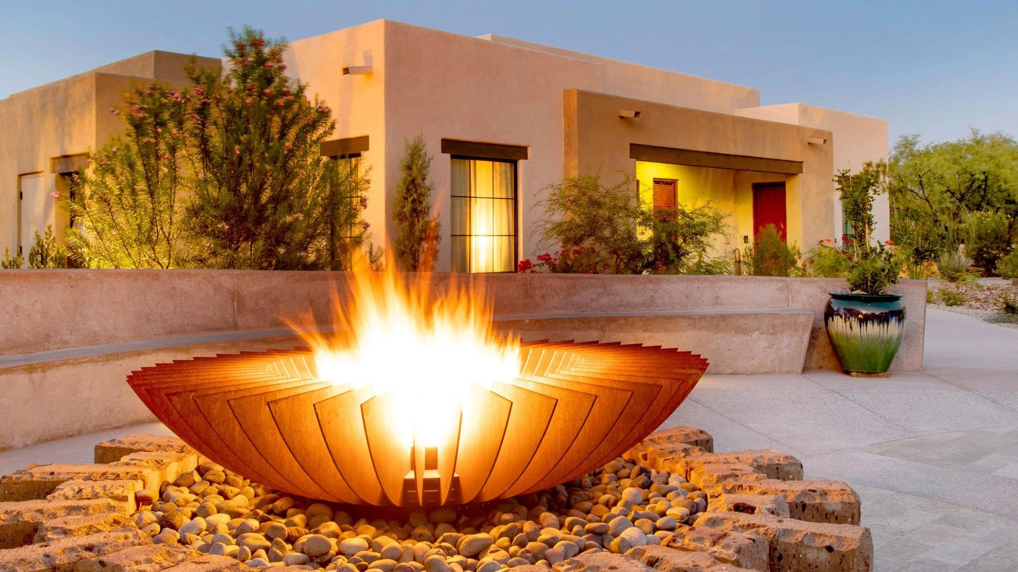 Modern house with desert landscaping illuminated by outdoor lighting and a fire feature in the foreground during dusk.