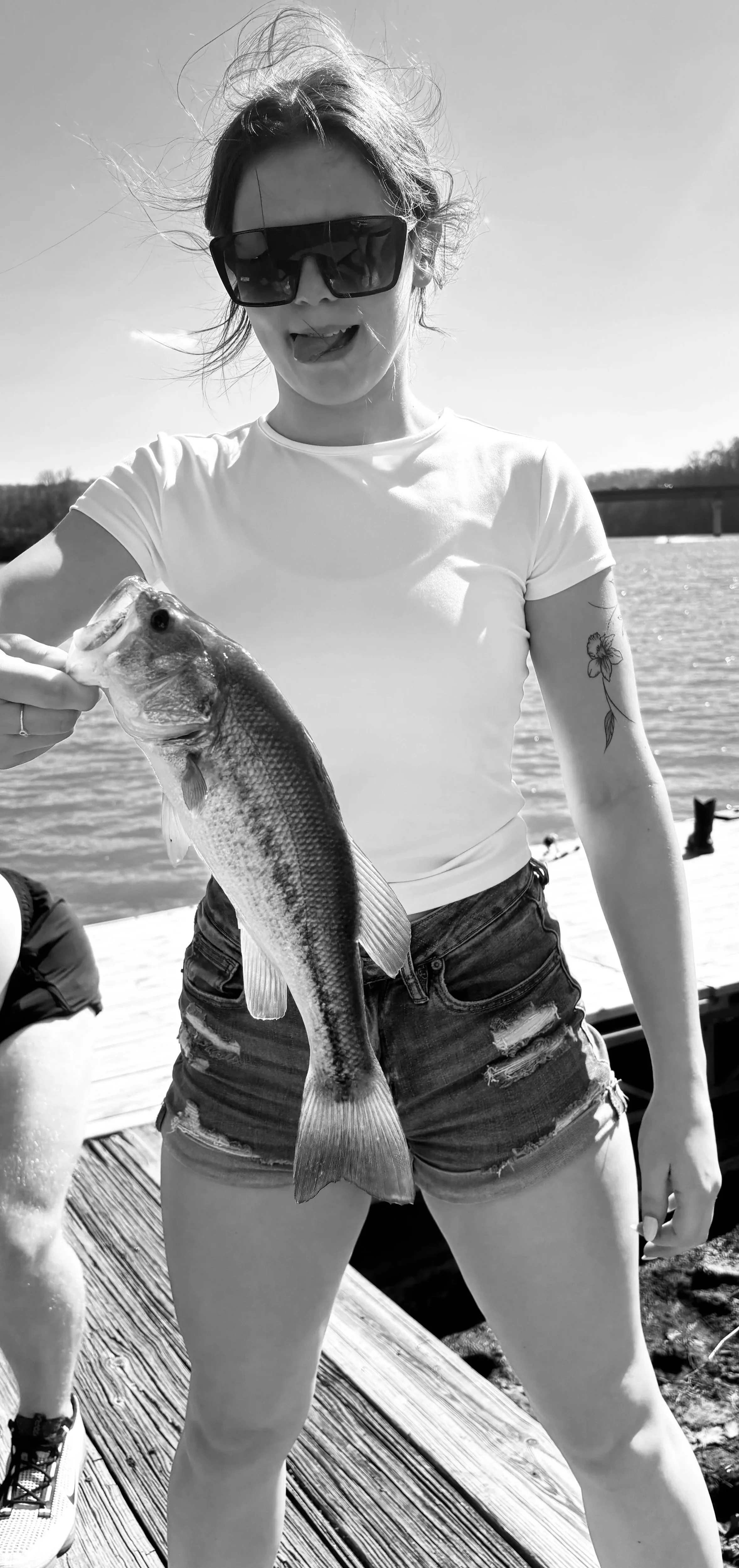 A woman wearing sunglasses and a white t-shirt, holding a fish she just caught, standing on a dock by a body of water.