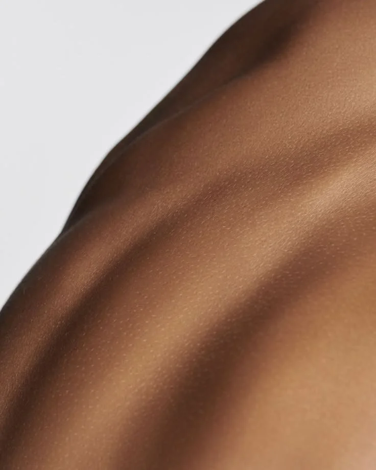 Close-up of smooth, brown skin, possibly a shoulder or arm, with subtle lighting highlighting the skin's texture.