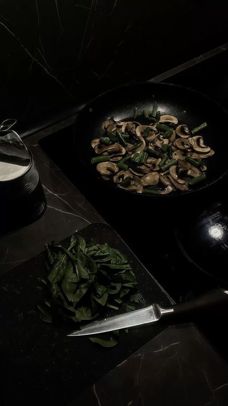 Sliced mushrooms and chopped green onions cooking in a black skillet, fresh spinach and a knife on a black countertop.