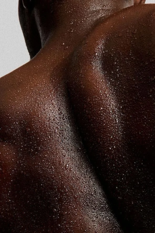 Close-up of a person's glistening dark skin with water droplets, focusing on shoulder and upper back area.