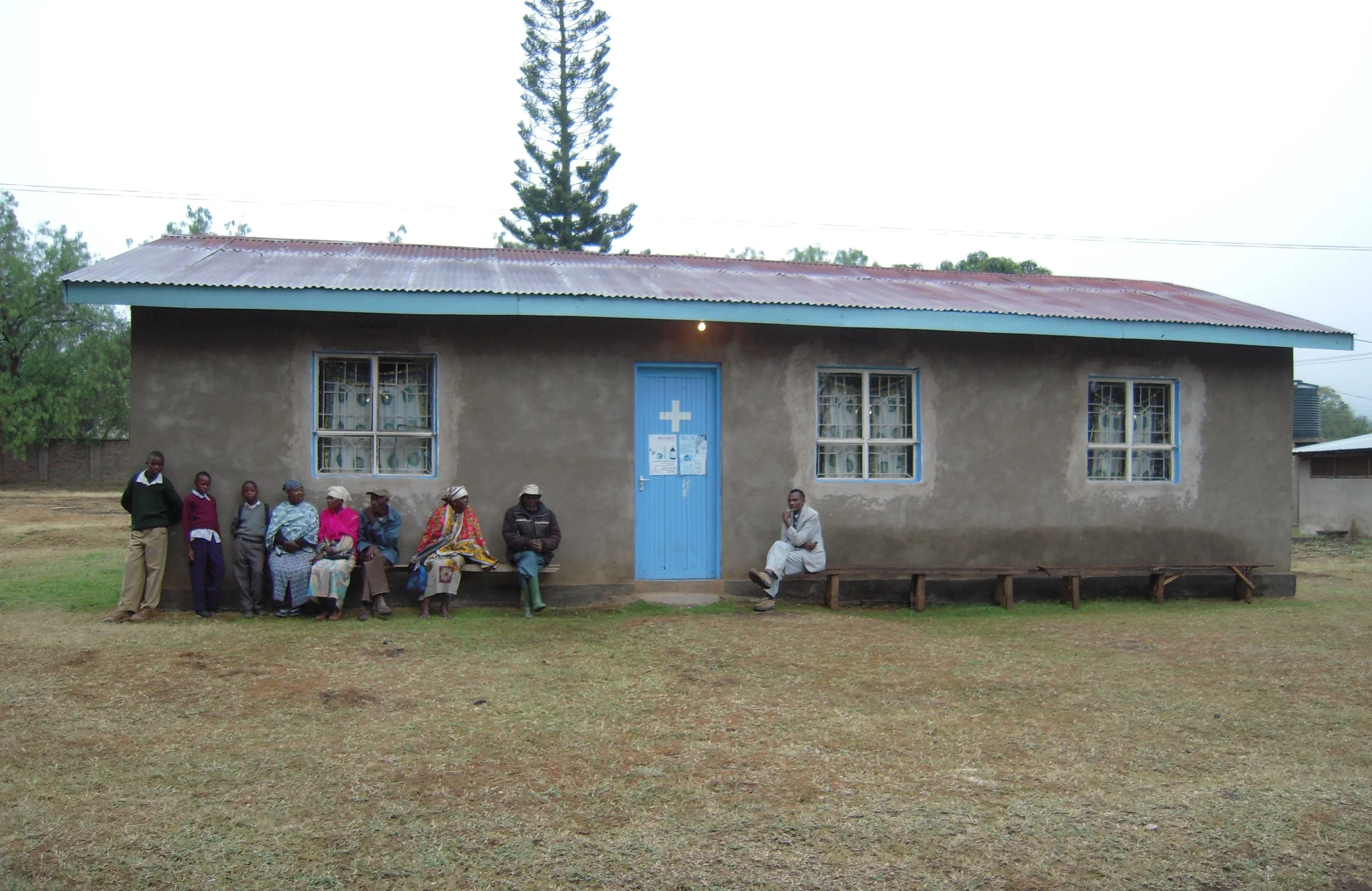 A simple single-story building with a blue door, three windows, and a corrugated metal roof. People are sitting and standing outside the building on a grassy area with a tree in the background.