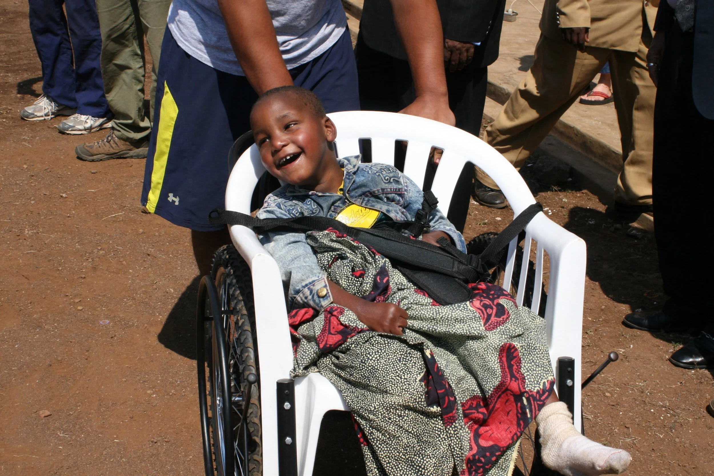 A young child sitting in a wheelchair outdoors, smiling, with several adults standing behind, some hands resting on the child's shoulders, on a dirt ground in a sunny environment.