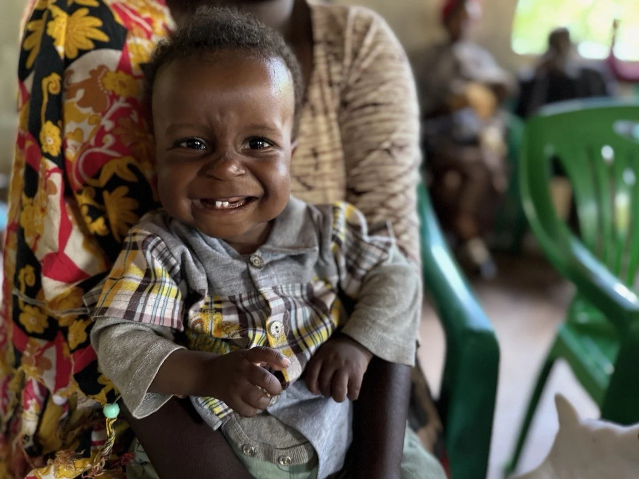 A smiling young African boy with missing front teeth, sitting on an adult's lap in a crowded room with green plastic chairs.