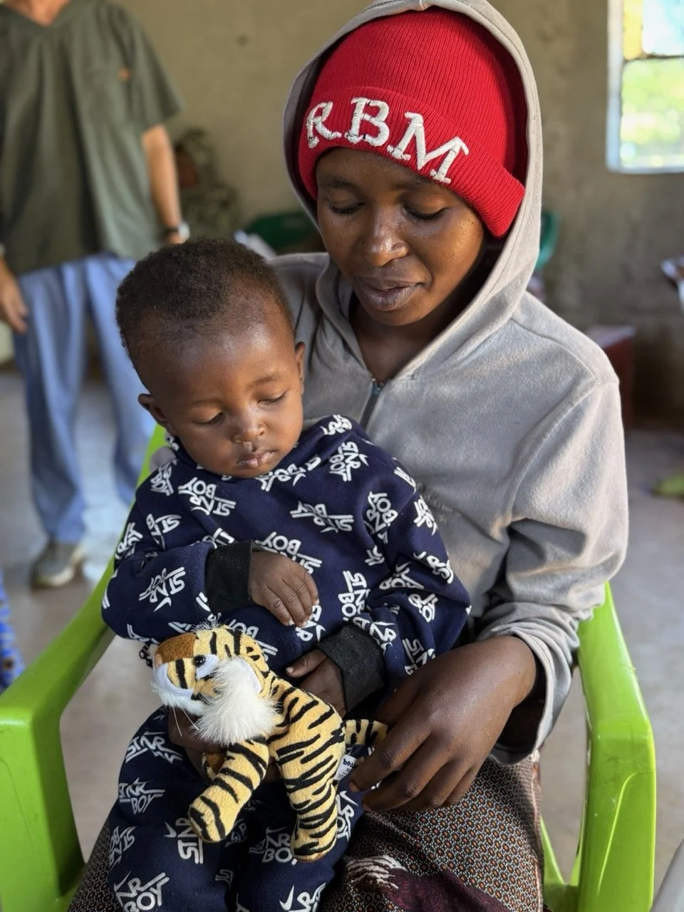 A woman holding a young boy sitting on her lap, both looking at a small stuffed tiger toy. The woman is wearing a gray hoodie and a red beanie with white letters, and the boy is in a navy sweatshirt with white text.