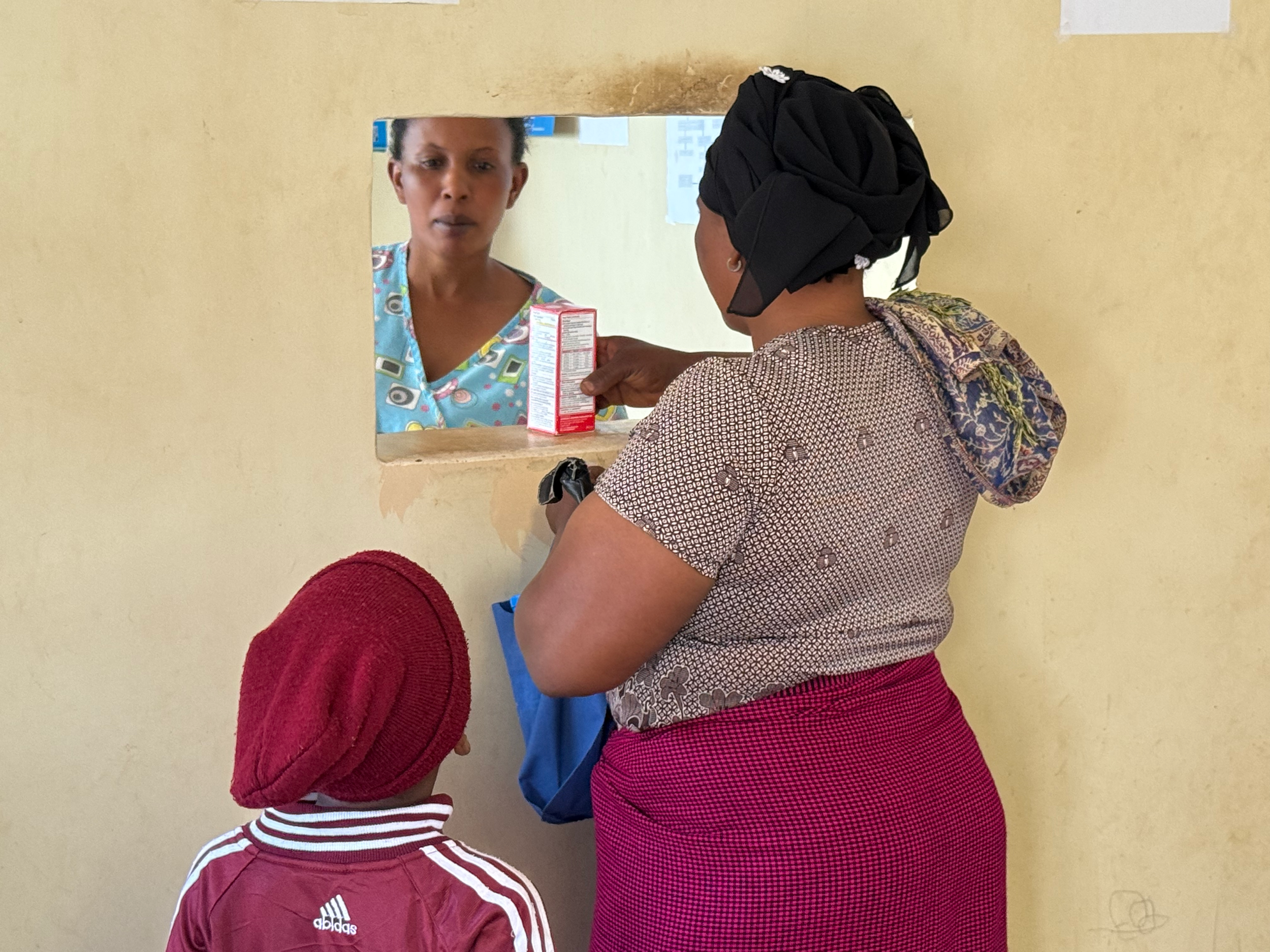 A woman with a black head covering and earrings is looking at her reflection in a mirror. In front of her, a small child wearing a maroon and white Adidas tracksuit is also looking at the mirror. The woman is holding a small rectangular object, possibly a medicine box, and the wall behind them is yellow with some papers attached.