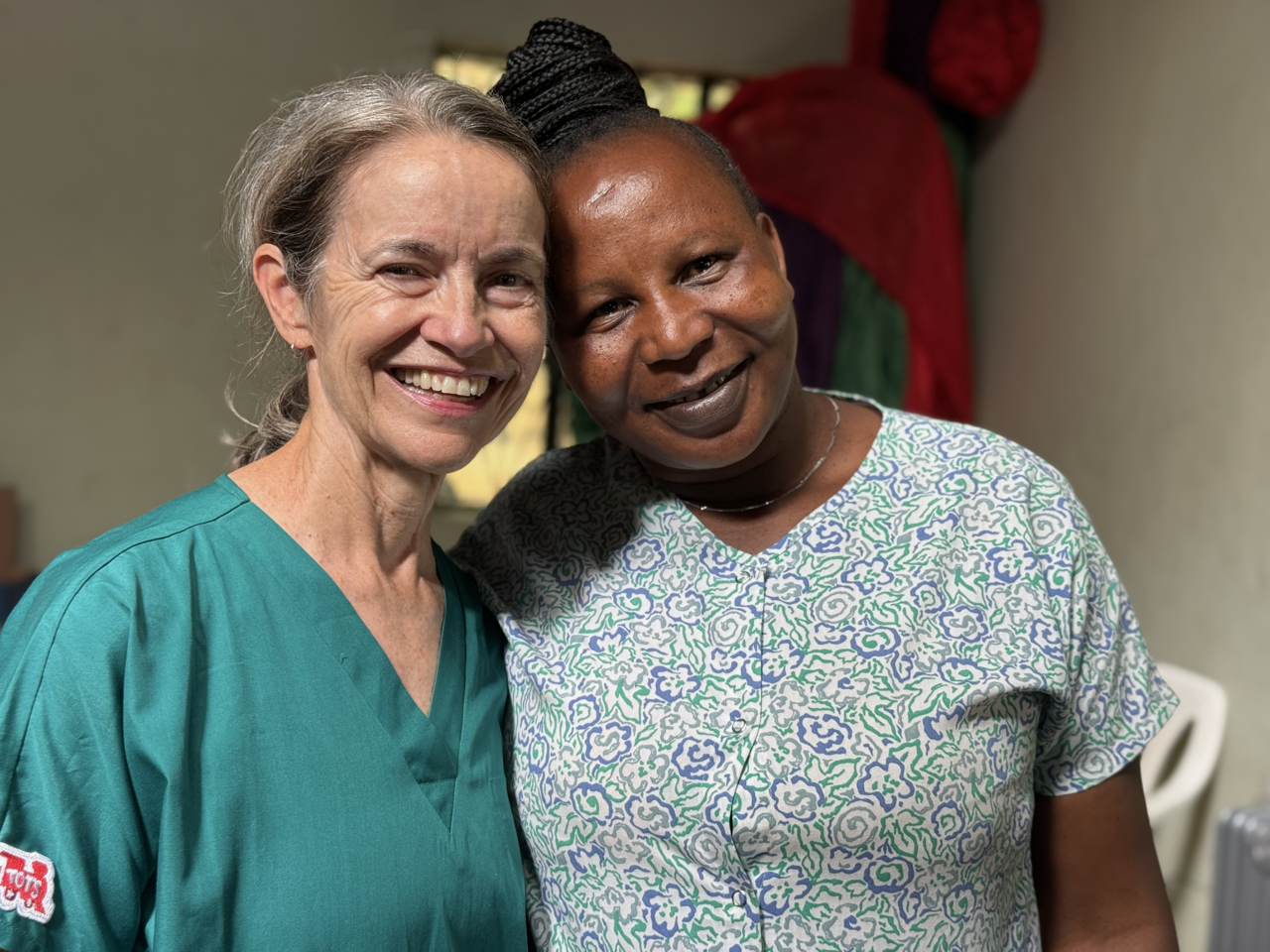 Two women smiling and posing together indoors, one wearing medical scrubs and the other in a patterned top.
