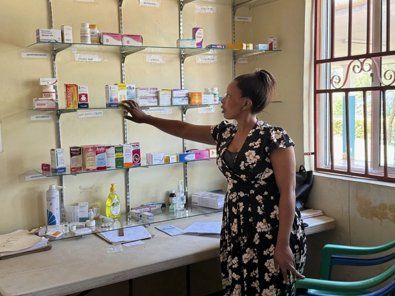 A woman in a floral dress reaching for medication on a cluttered pharmacy shelf, with a window to the right.