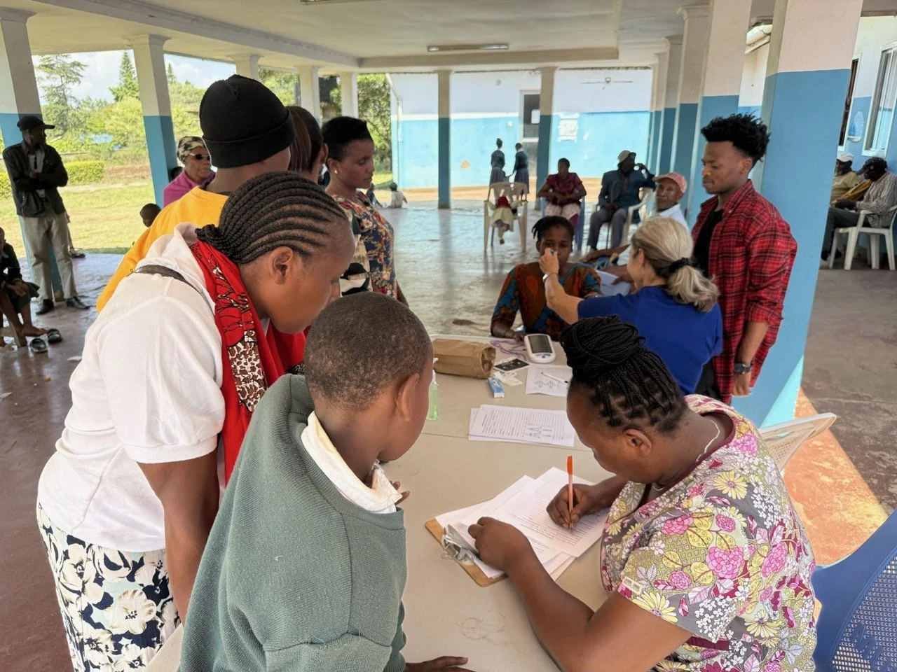 People filling out forms at a community health or registration event in a covered outdoor space with blue walls and a few chairs, with others sitting and standing in the background.