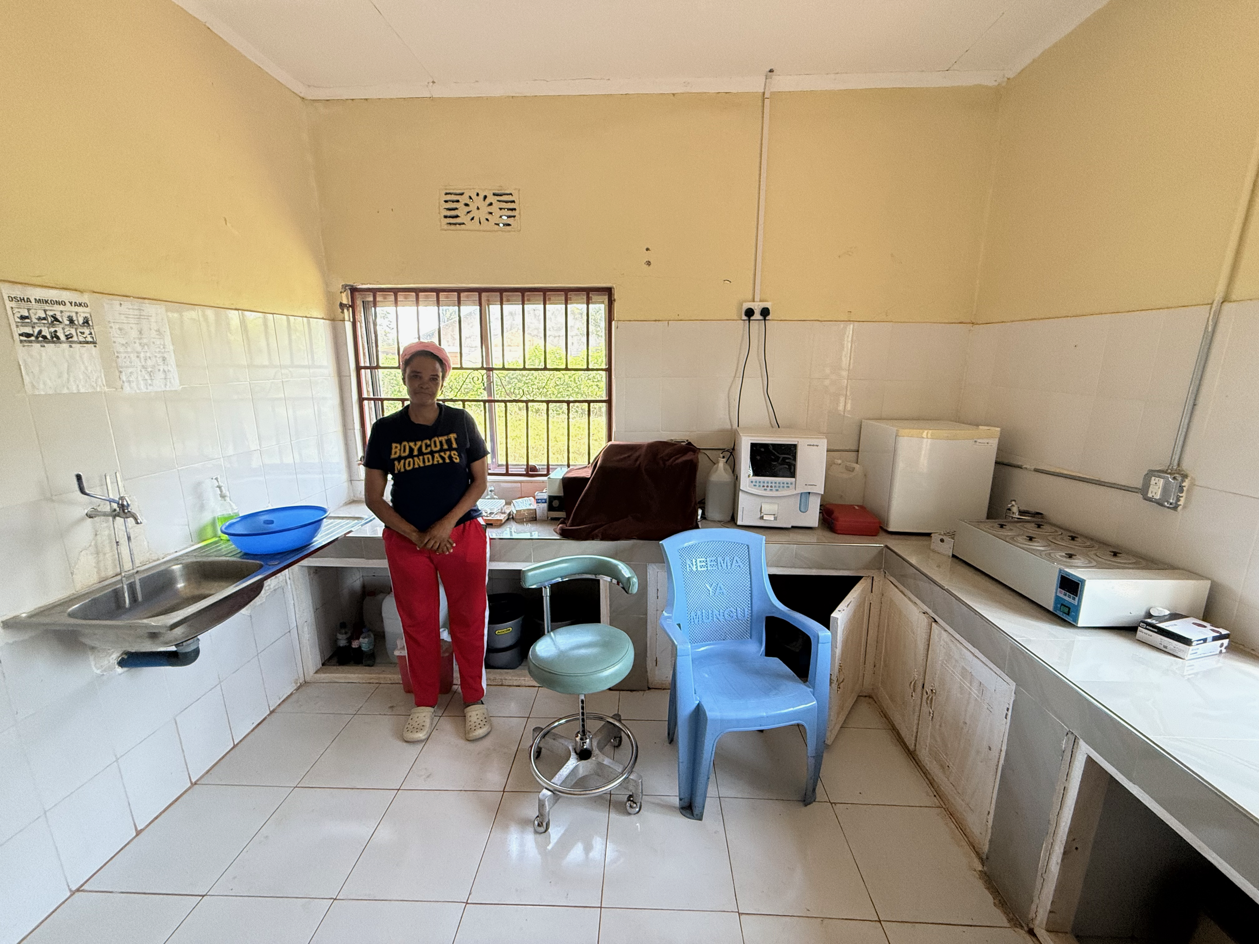 A woman standing in a kitchen with tiled walls and floor, wearing a black t-shirt with 'BOYCOTT MONDAYS' written on it and red pants. The kitchen has a blue chair, a sink with a blue basin, and various appliances and items on the counter. There's a window behind her with bars, and the ceiling has an air vent.