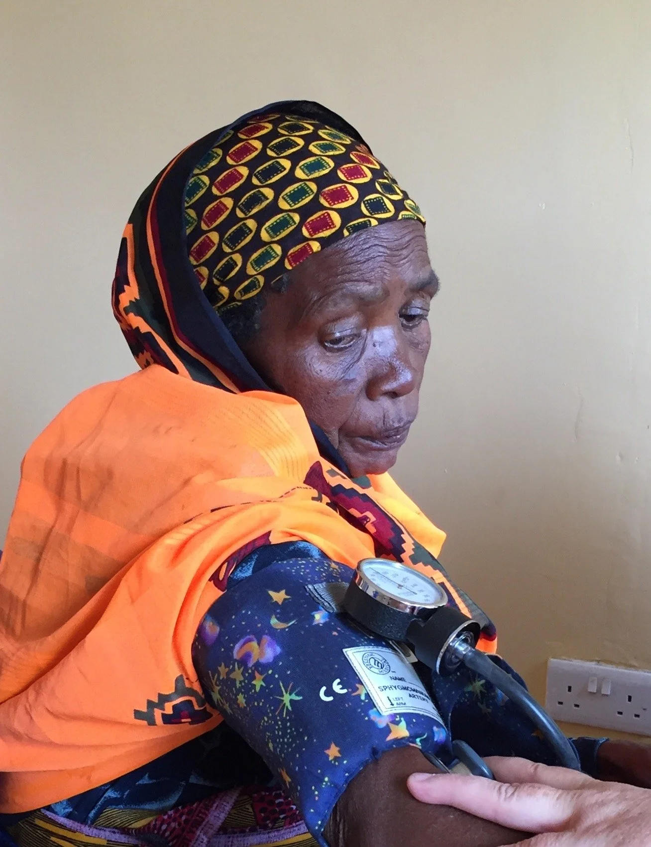 An elderly woman with dark skin and gray hair, wearing a colorful headscarf and bright orange clothing, receiving a medical blood pressure check from a healthcare professional.
