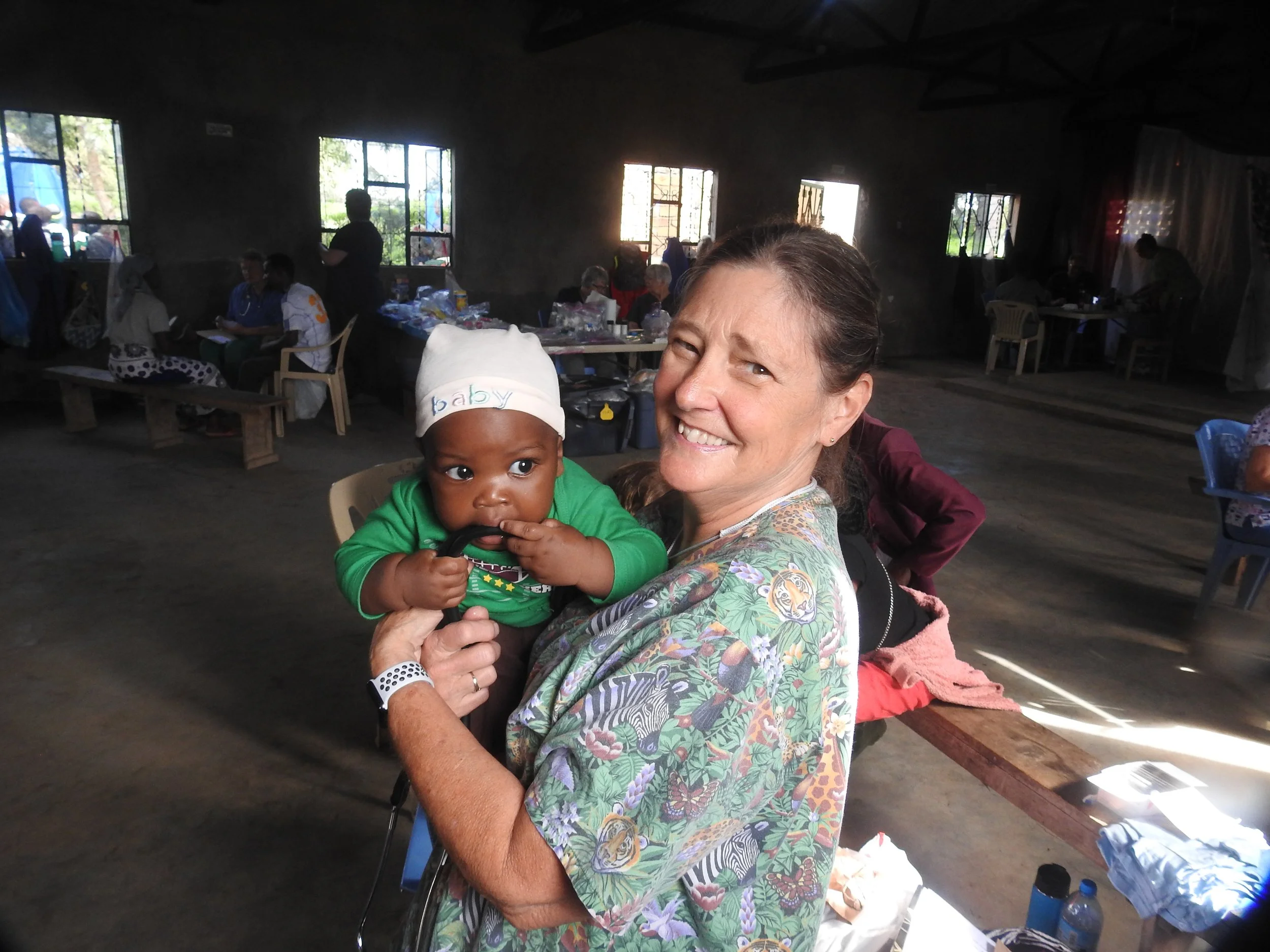 A woman holding a baby with a hat that says 'baby' on it, inside a dimly lit community hall or gathering space with other people at tables in the background.