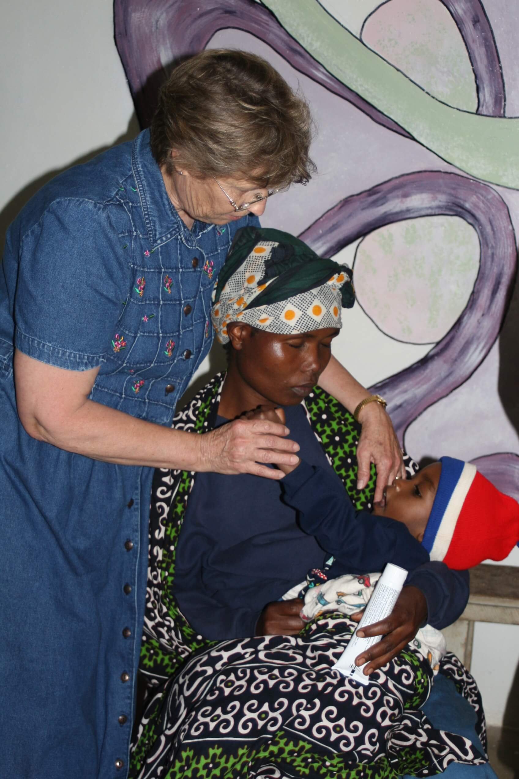 A woman appears to be giving a health check or consultation to a young man. The woman is leaning over and placing her hand on his shoulder, while the young man is sitting and holding a bottle and a pamphlet or piece of paper. In the background, there is abstract wall art with purple, green, and black shapes.
