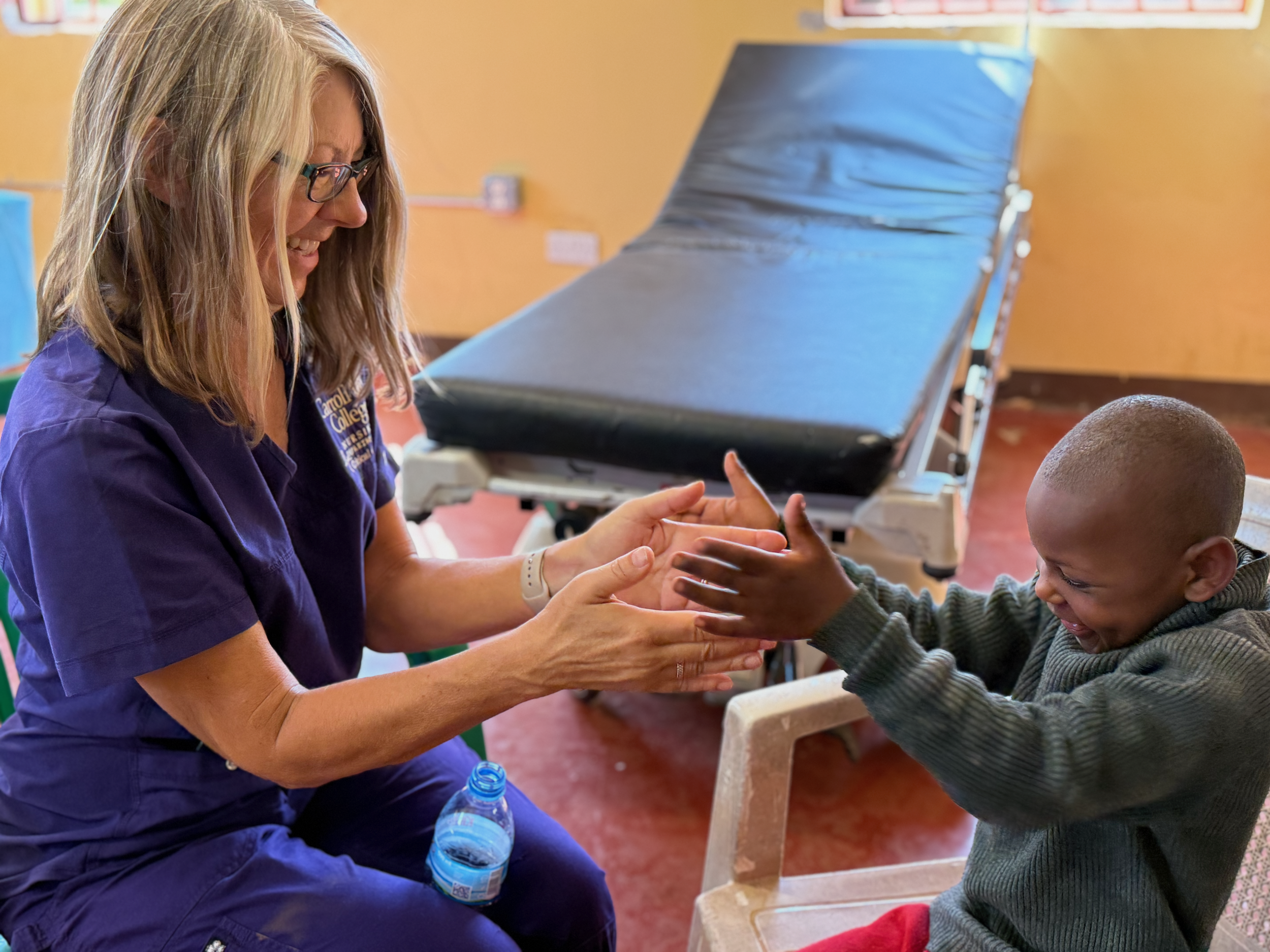 A woman with blonde hair, glasses, and a purple shirt is sitting on a chair, smiling and holding hands with a young boy. The boy has dark skin, short hair, and is wearing a gray striped sweater. They are interacting happily in what appears to be a medical or therapy setting, with a hospital bed or examination bed in the background.
