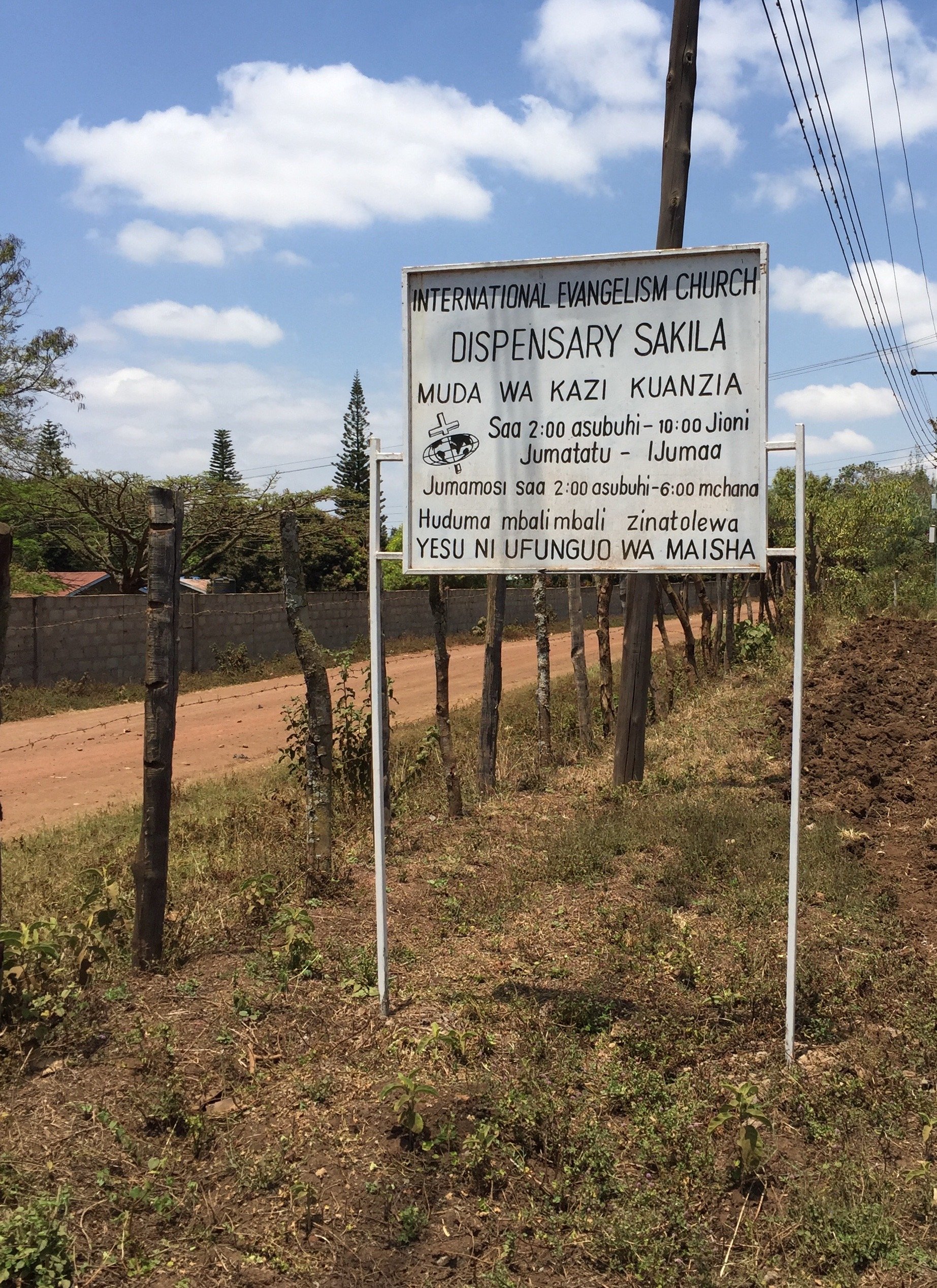 Signboard for International Evangelism Church and dispensary Sakila with service hours and additional text in Swahili, located outdoors near a dirt path with trees and a fence in the background.