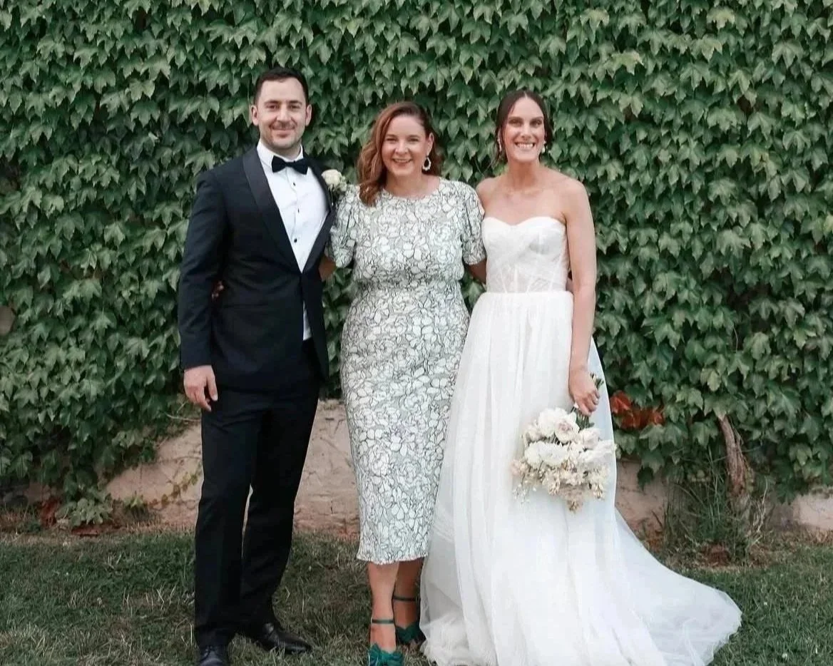 Married by Beck - Melbourne and Yarra Valley celebrant is standing in front of a green ivy wall at Stones of The Yarra Valley with a bride and a groom. The groom is wearing a tuxedo and the bride is wearing a white dress and holding a bouquet.