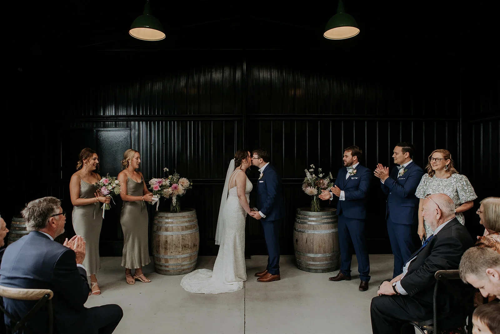 A wedding ceremony with a bride and groom kissing, surrounded by bridesmaids and groomsmen, in front of an audience, with flower arrangements on barrels and decorated with pink and white flowers.
