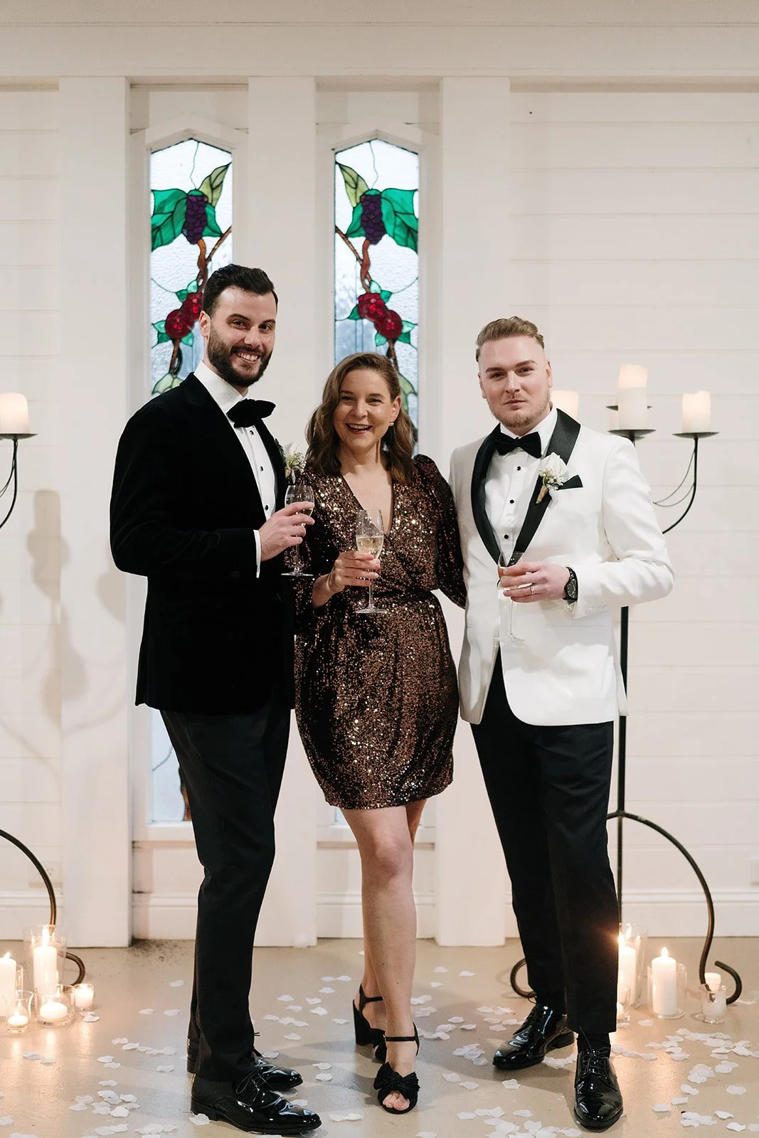 Three people at a wedding celebration standing in front of stained glass windows, holding glasses of champagne. The two men are dressed in tuxedos, and the woman is wearing a sparkly dress. Candles and flower petals decorate the floor.