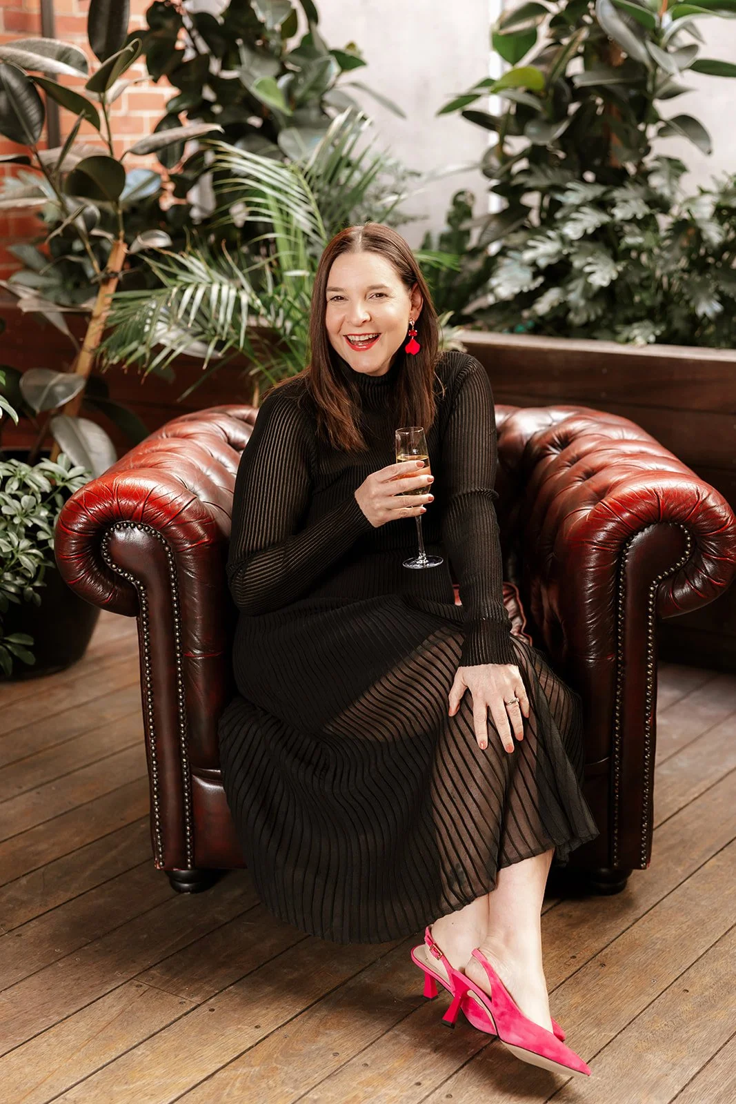 A woman in a black dress with pink high heels and red earrings sitting on a leather sofa, holding a glass of champagne, surrounded by lush green plants.