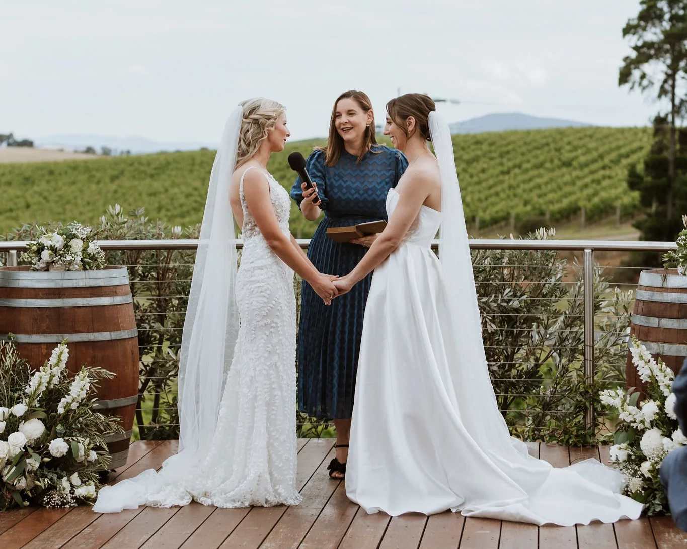 Two women in wedding dresses holding hands during a wedding ceremony outdoors on a wooden deck with wine barrels and floral arrangements, officiant in blue dress, green hills in background.