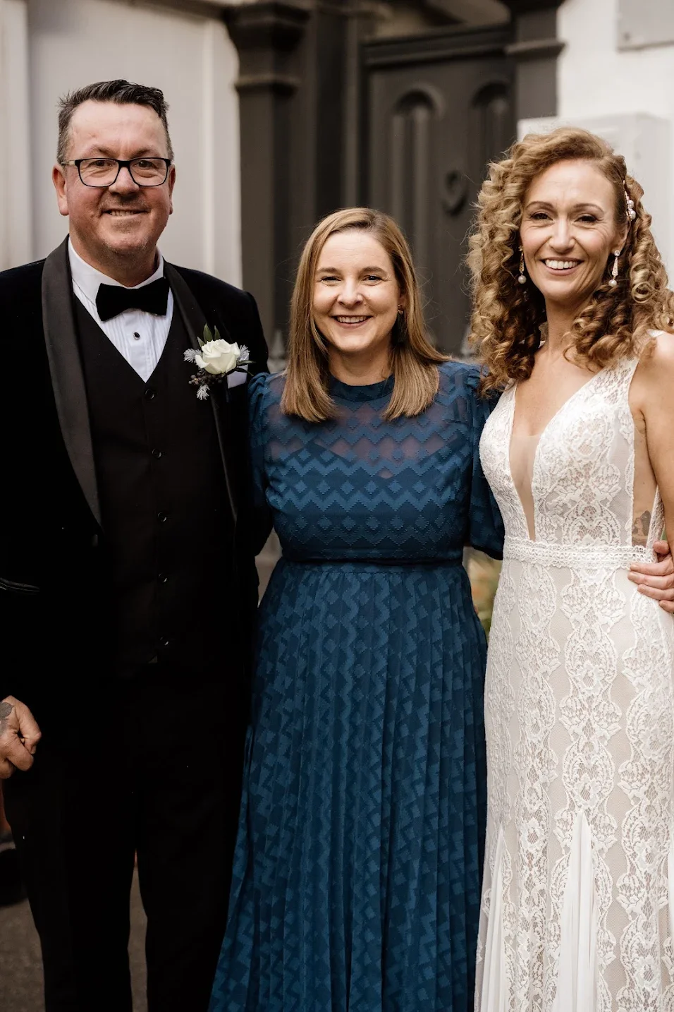 Three people smiling at a formal event, with a man in a black tuxedo, a woman in a blue dress, and a woman in a white lace dress, standing close together indoors.