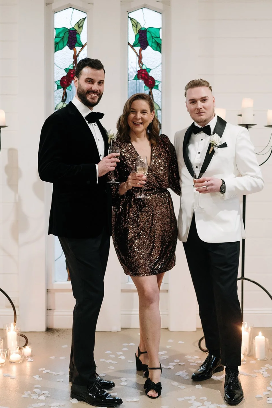 Three people dressed in formal attire celebrating with glasses of champagne inside a decorated venue with stained glass windows and candles.
