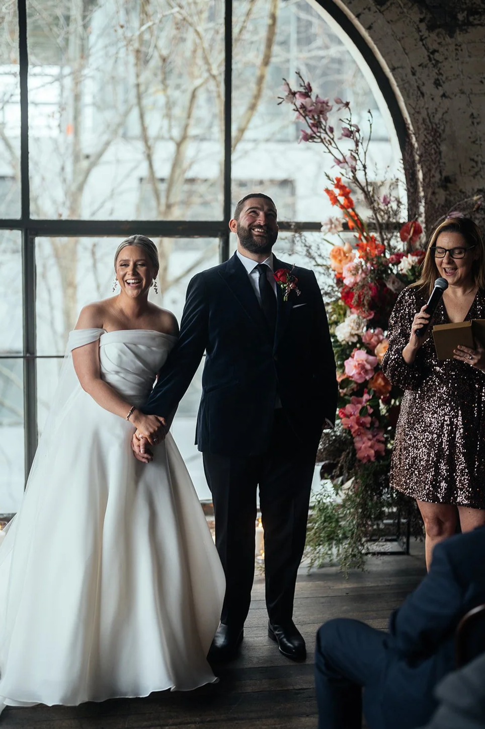 A smiling bride and groom holding hands at their wedding ceremony, with a woman officiant reading from a book and speaking into a microphone, in front of a large window decorated with flower arrangements.