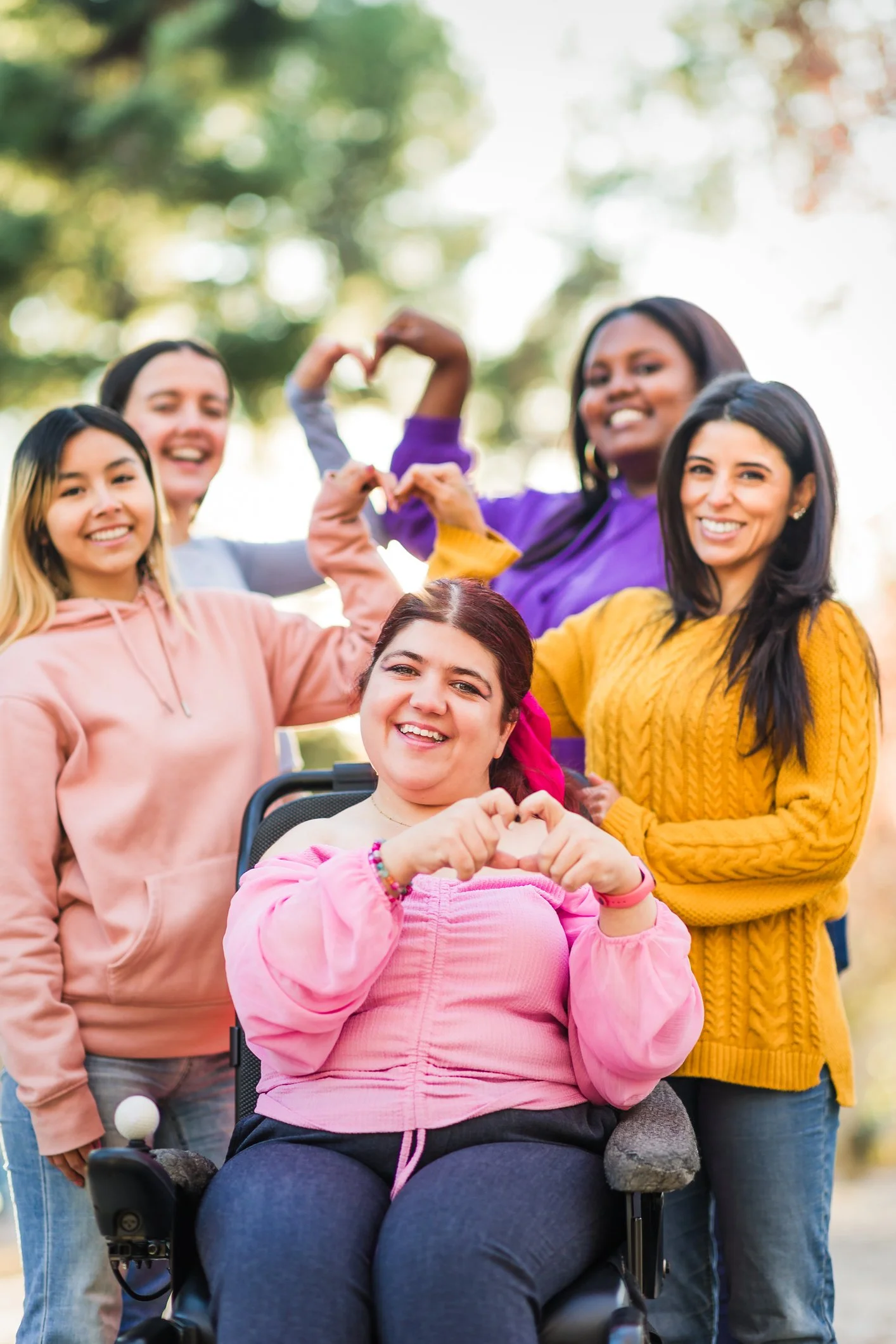 A group of five diverse women outdoors, smiling and making heart shapes with their hands, with one woman in a wheelchair in the front.