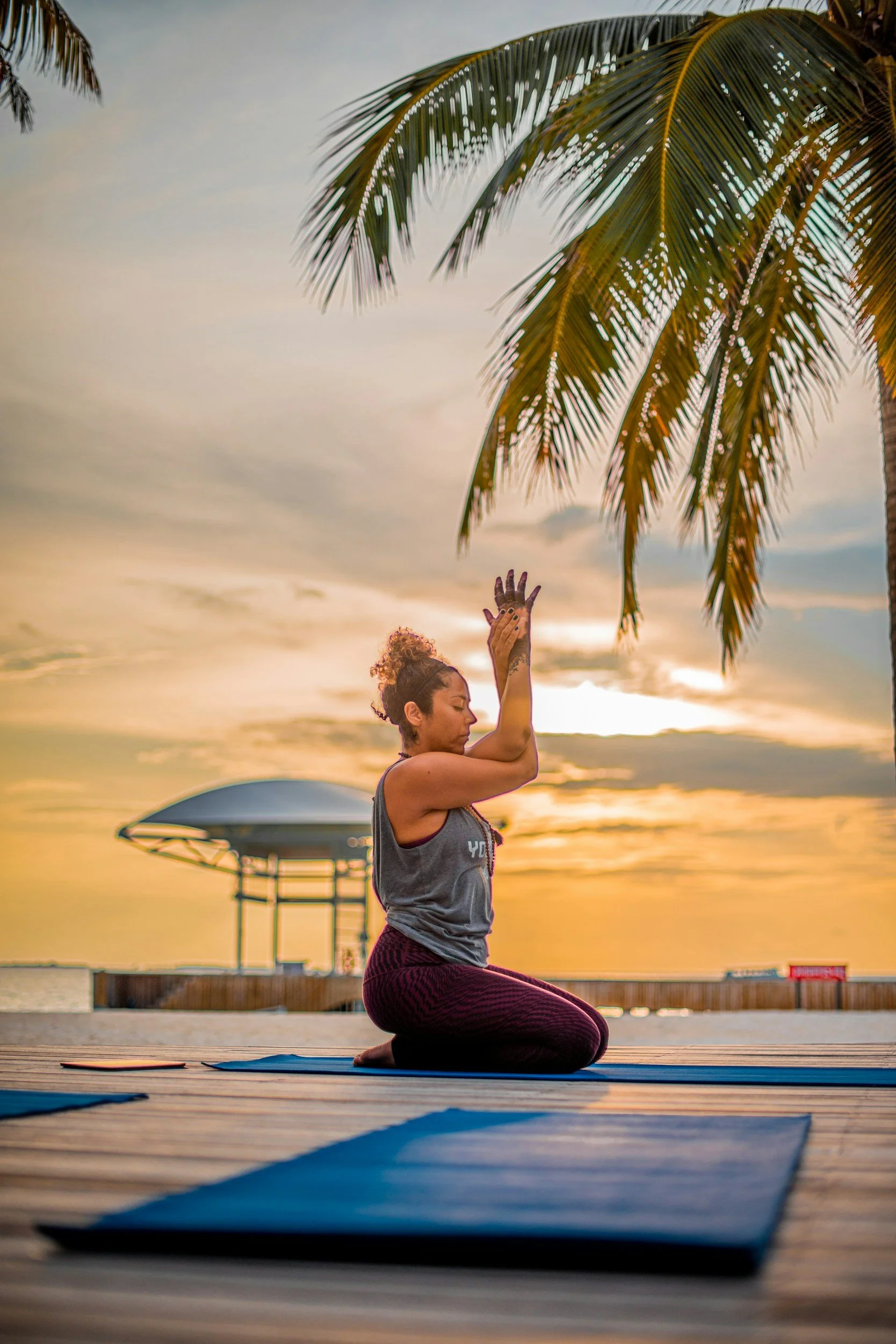 A woman practicing yoga on an outdoor deck at sunset with palm trees and a lifeguard tower in the background.