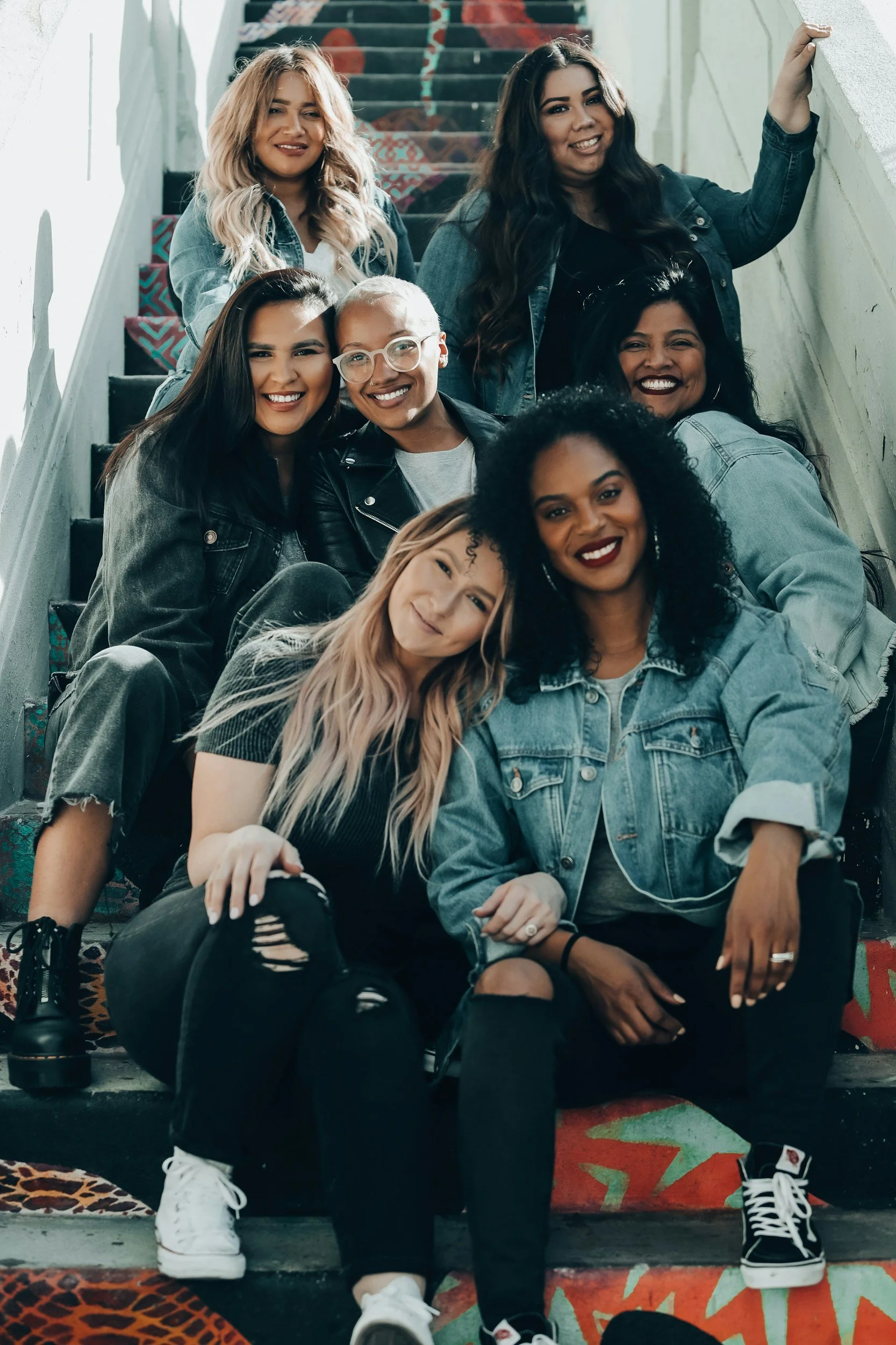 A group of nine diverse women sitting on a colorful staircase, smiling and looking at the camera, dressed in casual denim and black clothing.