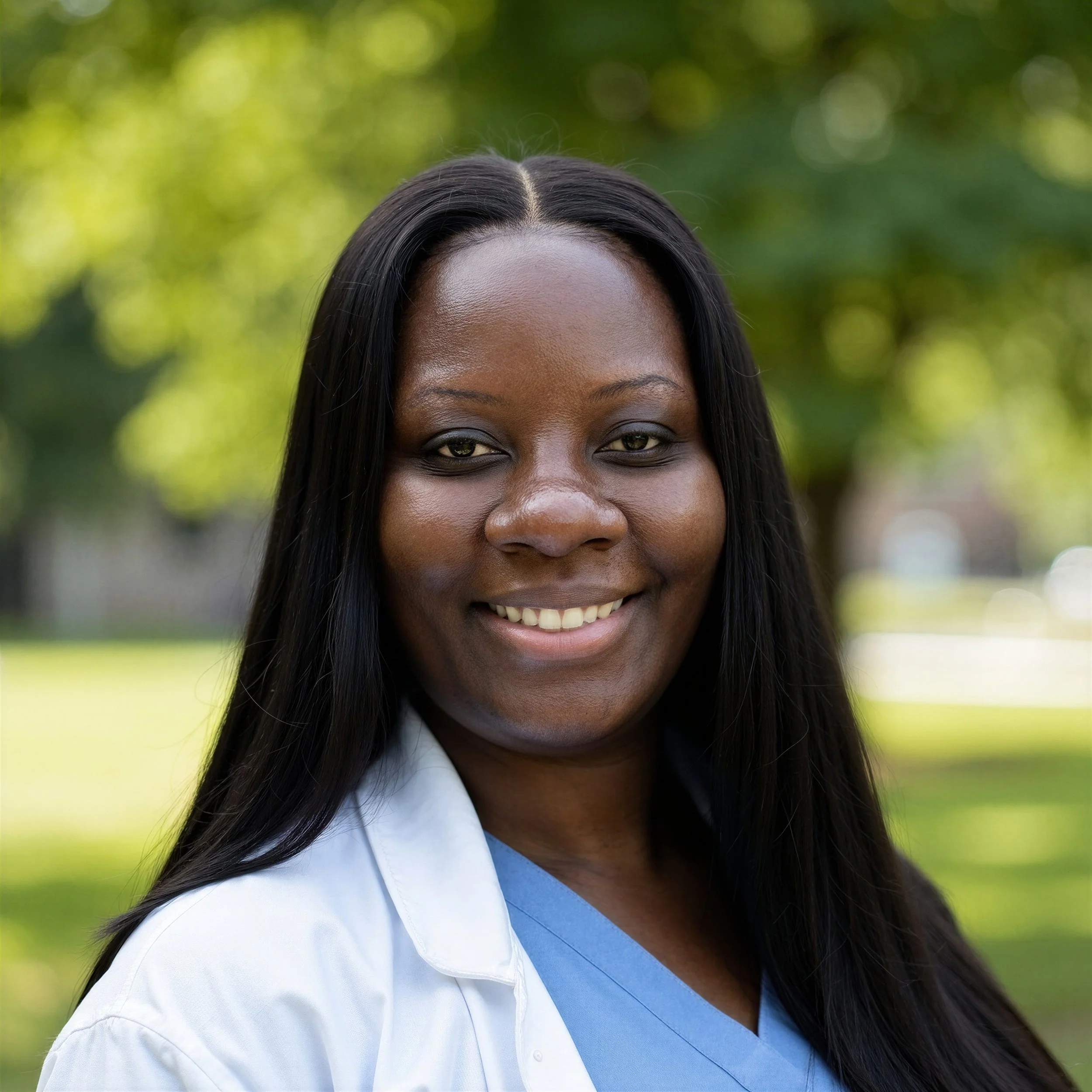 A smiling Black woman with long black hair, wearing a white coat and light blue scrubs, outdoors with blurred green trees in the background.